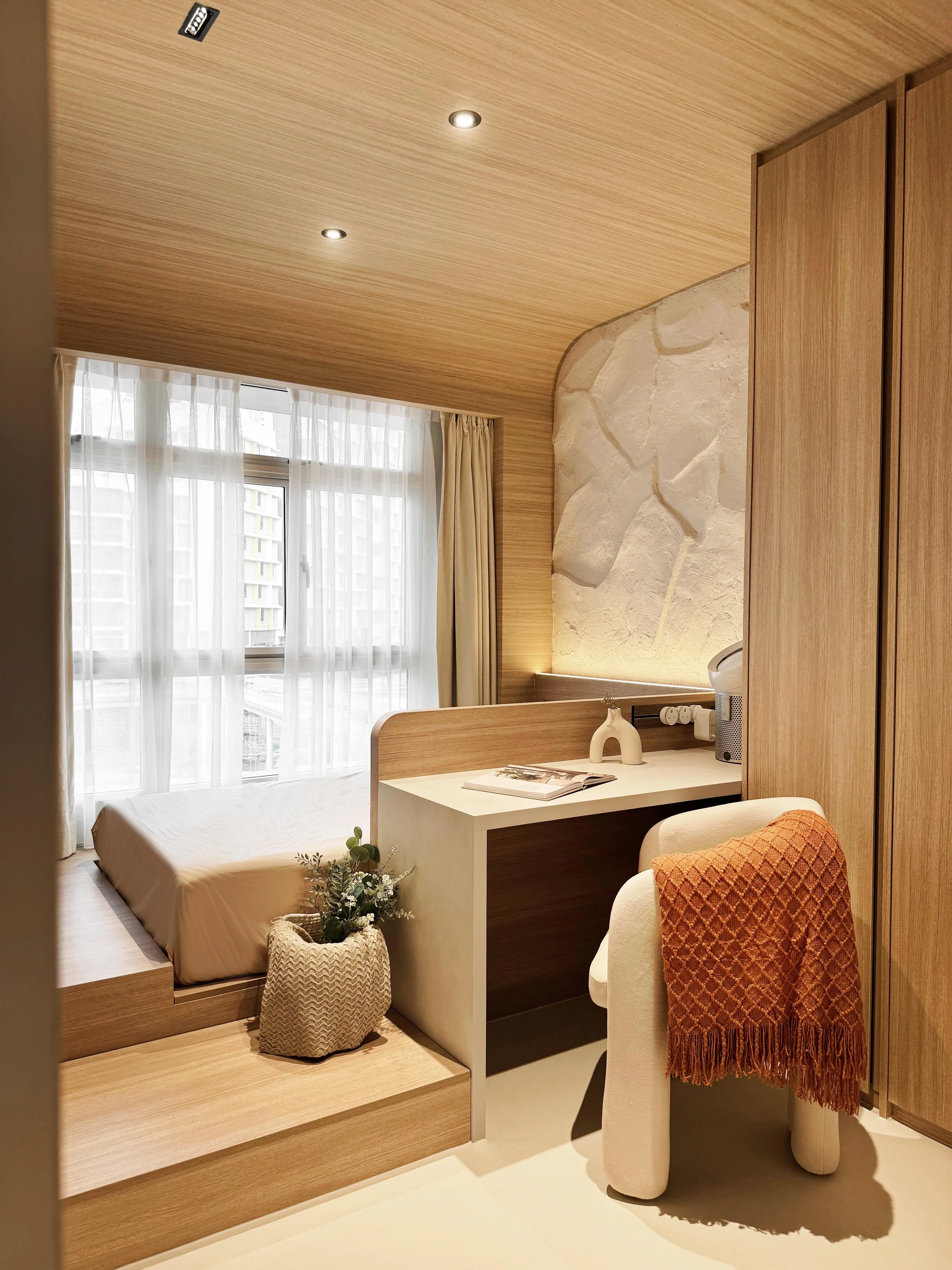 A cozy hotel room with a bed, a window with sheer curtains, a small desk with a chair and a decorative red blanket, a plant in a textured pot, and a wall with textured stone decor.