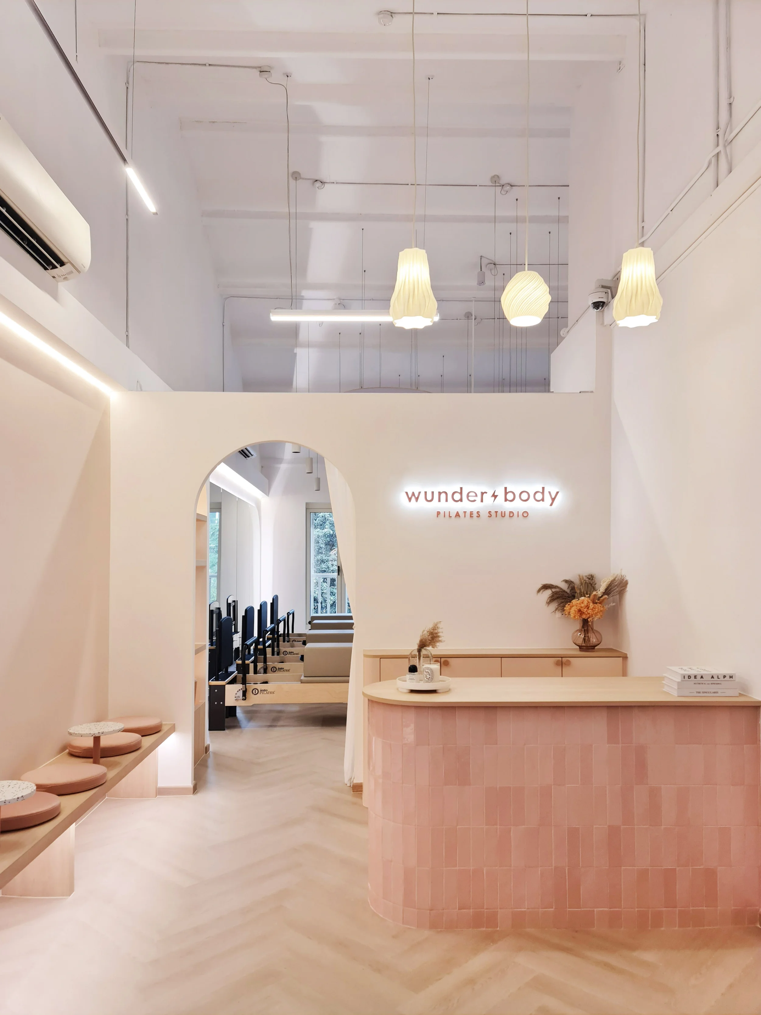 The interior of a Pilates studio with a reception desk, pink tiled counter, modern hanging light fixtures, and a waiting area with seating cushions. The studio has a minimalistic, pastel color palette with natural light coming through windows and fitness equipment visible in the background.