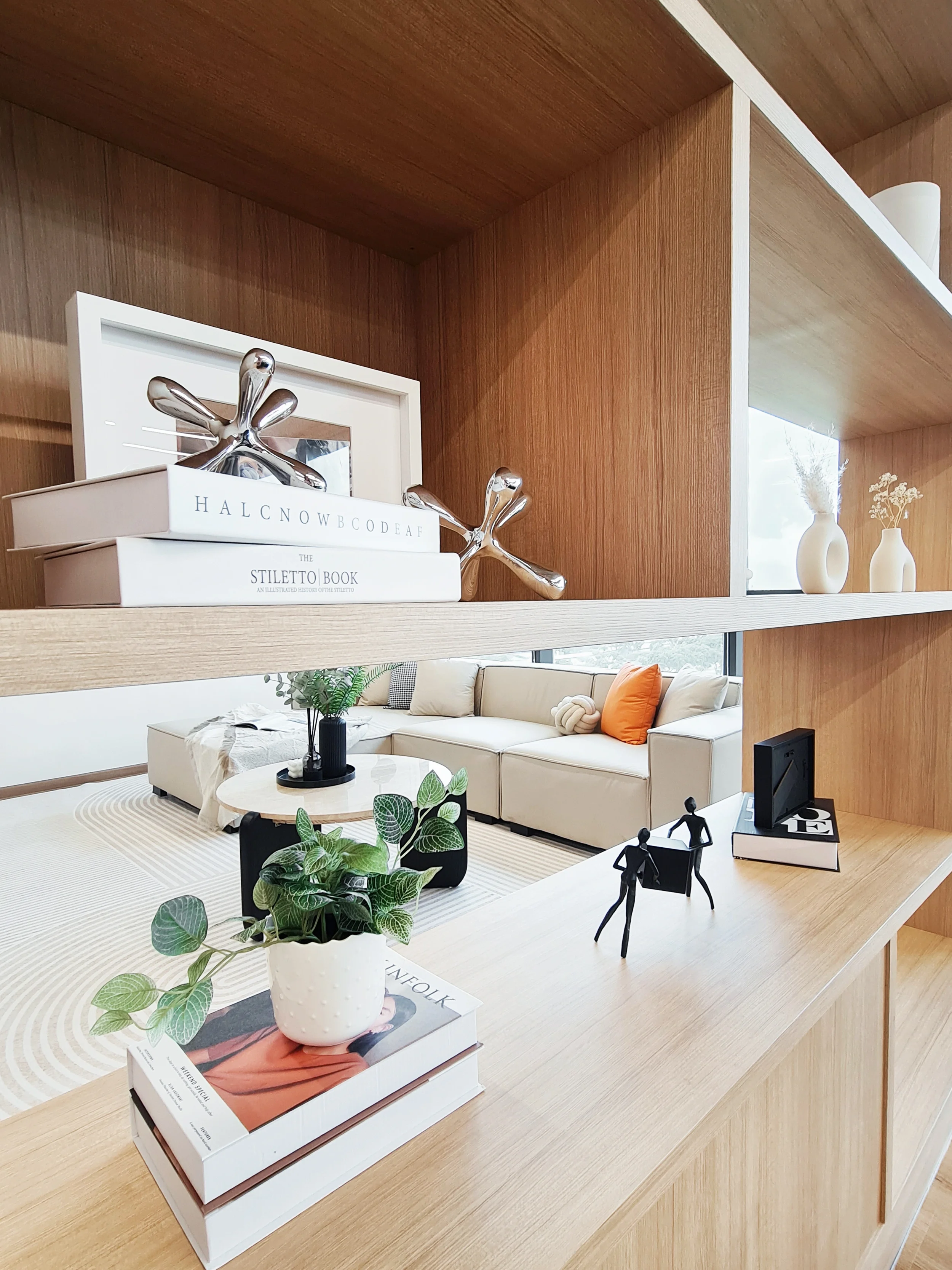 Decorative objects on wooden shelves in a modern living room, including books, metallic art sculptures, vases, and small figurines, overlooking a beige sofa with cushions and a coffee table with plants.