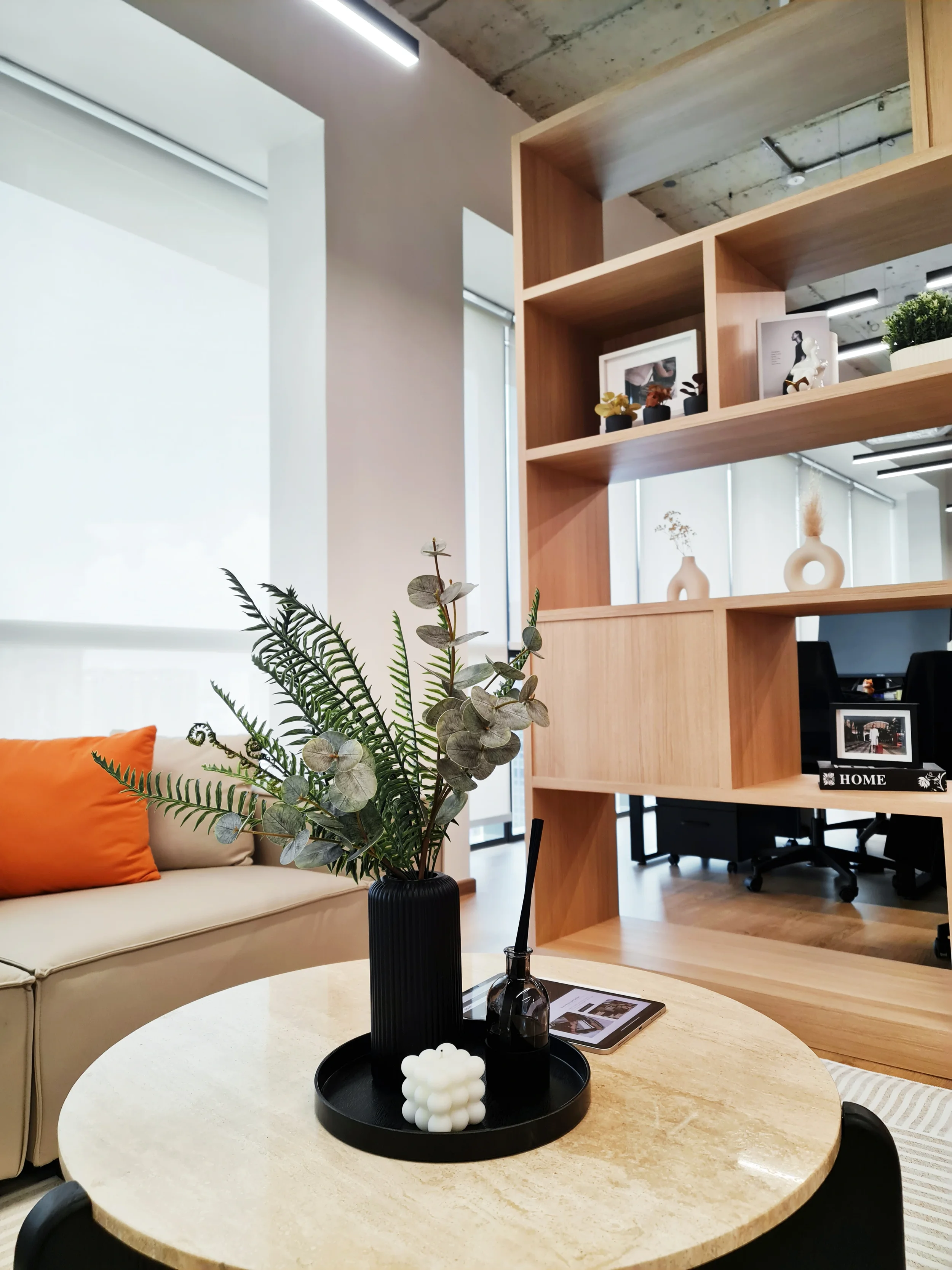 Modern office space with a round table decorated with a black tray holding a flower vase with greenery, a candle, and a perfume bottle. In the background, a beige couch with an orange pillow fronted by large windows, and a wooden bookshelf with decorative items and framed photos.