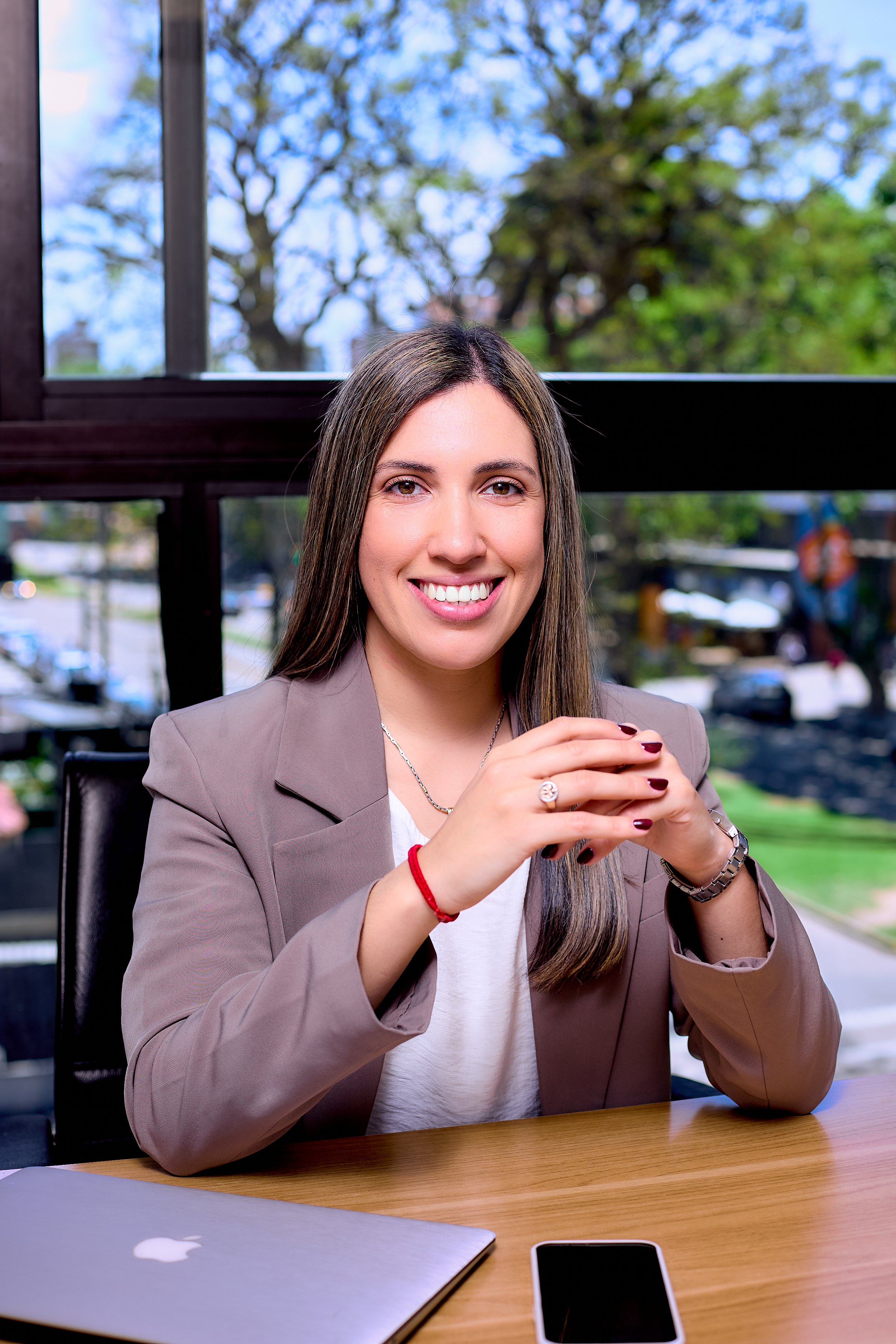 Mujer sonriendo en una oficina, con una laptop, teléfono móvil y ventanas con vista a árboles y calles.