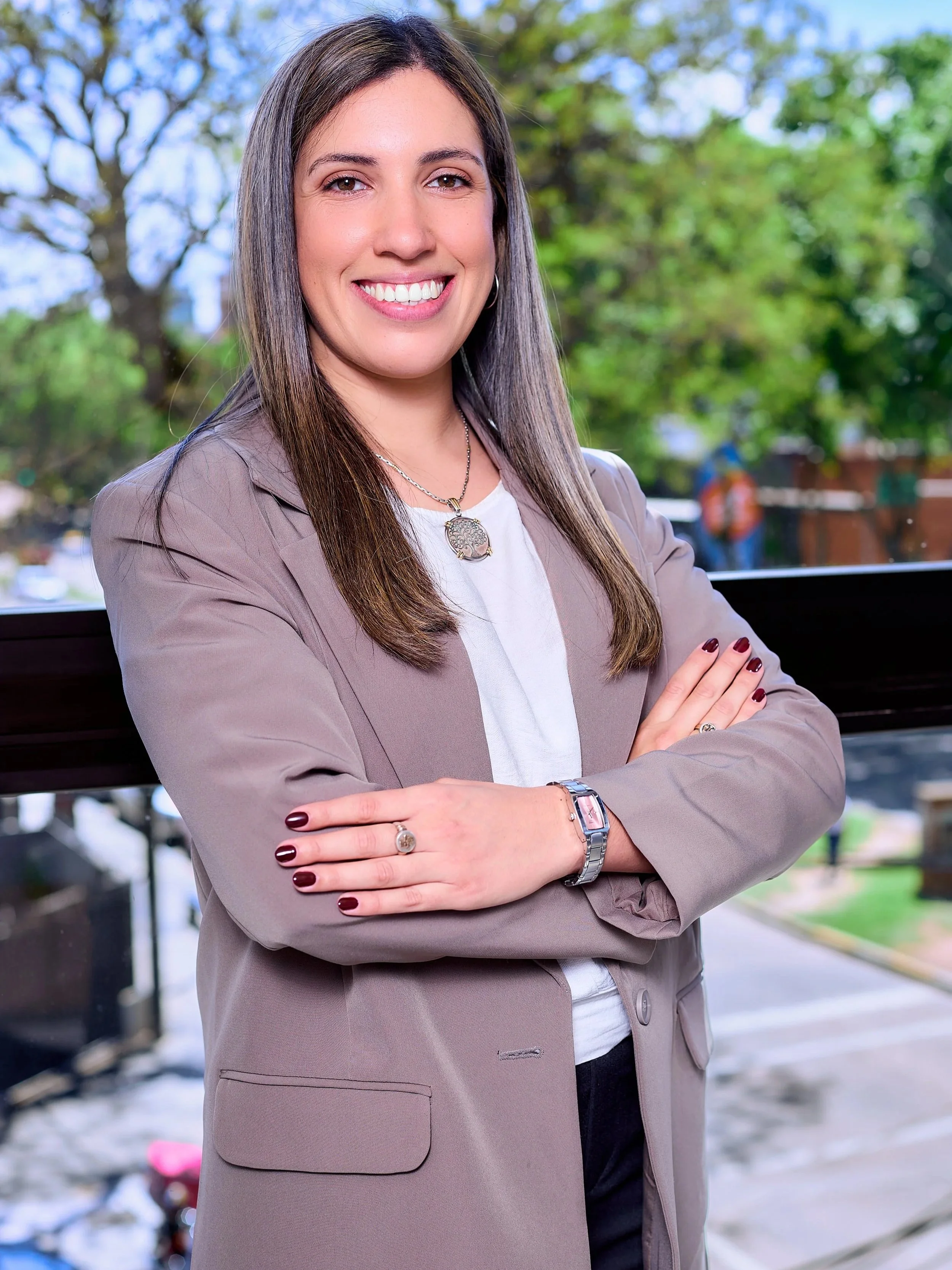 Mujer sonriendo con brazos cruzados, vista de frente, vestida con blusa blanca y saco gris, fondo de árboles y parque.