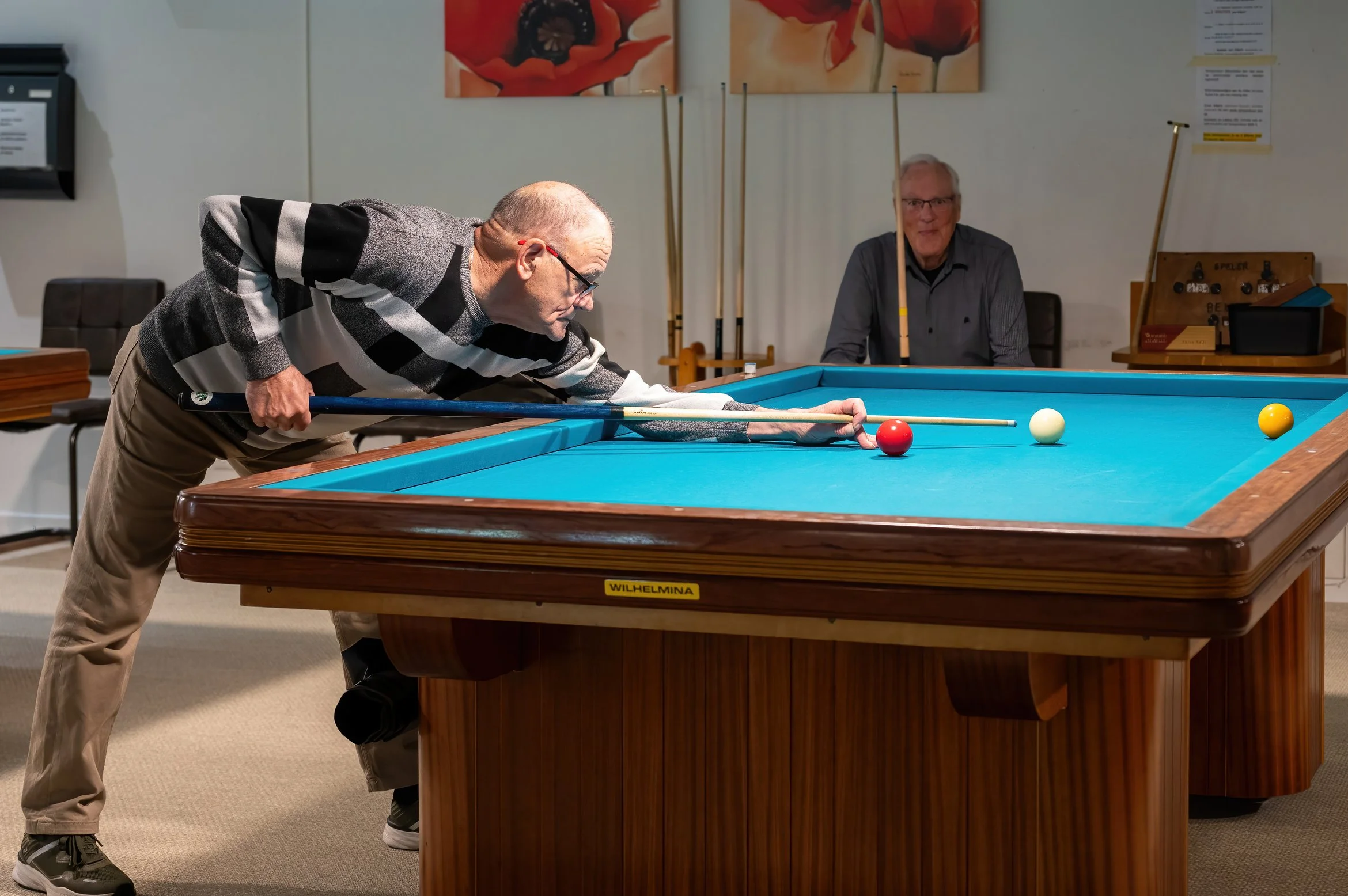 Two senior men playing a game of billiards indoors, with one leaning over aiming his shot and the other watching from behind on a blue felt table.