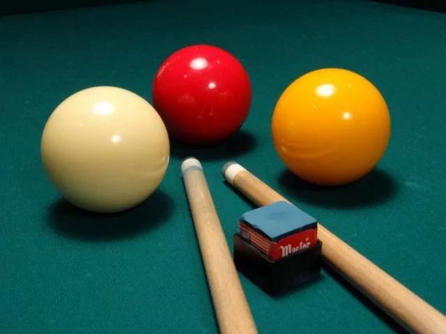 Three billiard balls (white, red, and yellow) on a green pool table, with two cue sticks and a box of chalk.