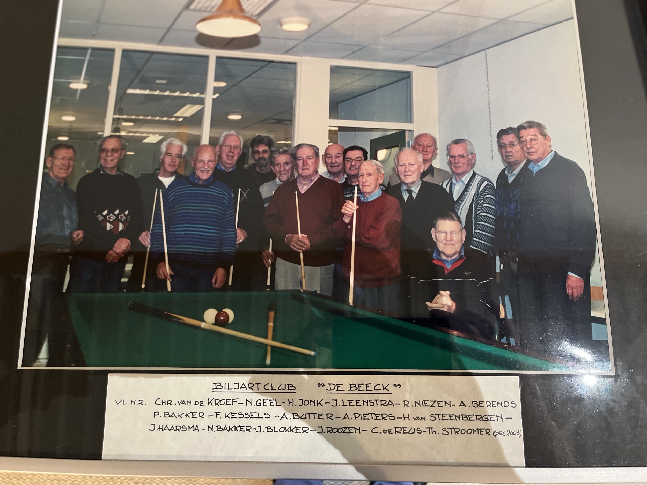 Group of men gathered around a billiard table, some holding pool cues, in a room with glass partition walls. There is a label identifying them as the Biljart Club 'De Beeck' with a list of names.