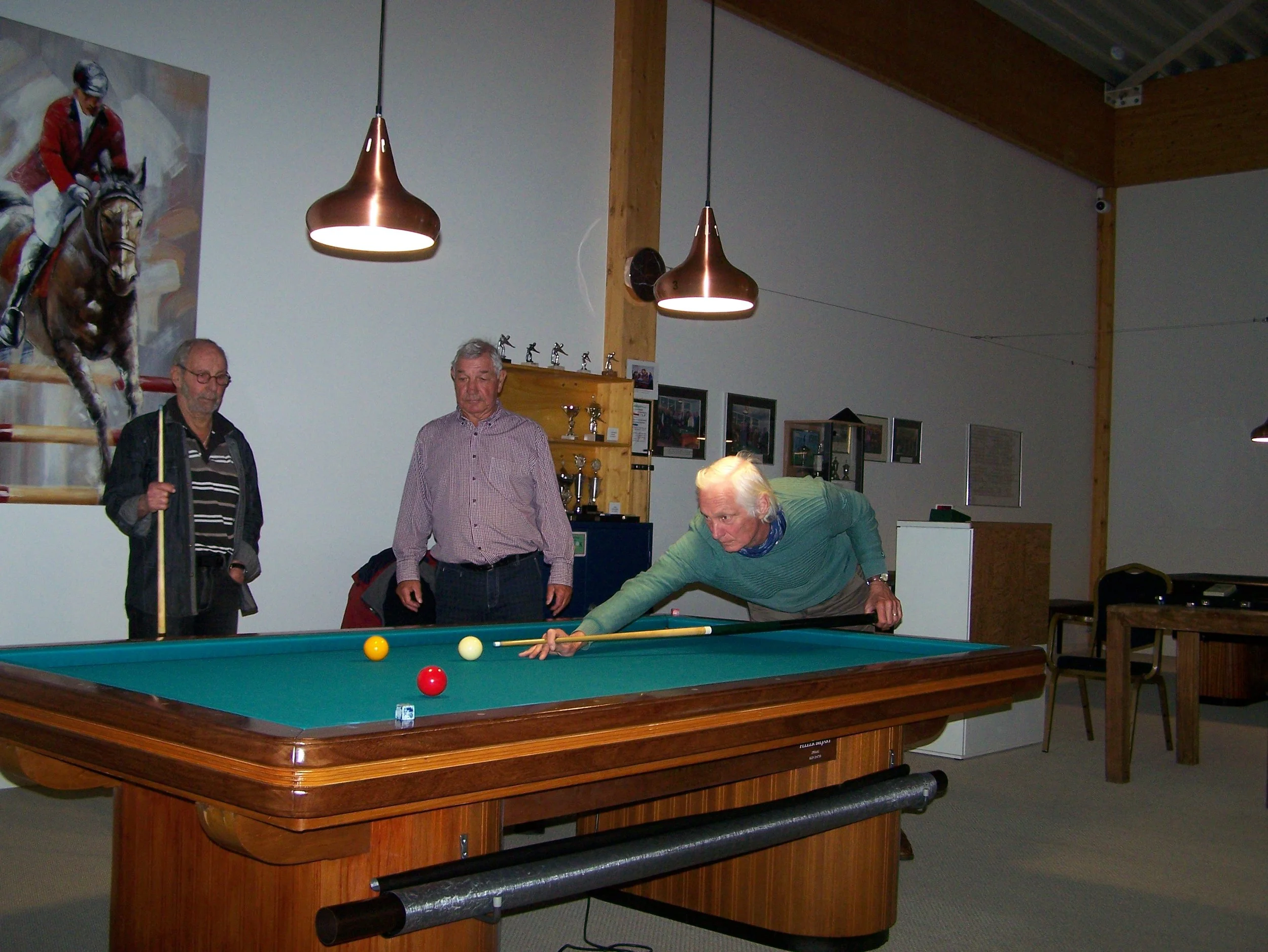 An elderly man playing pool on a green felt billiards table, aiming to hit the cue ball with a cue stick, while three onlookers watch him.