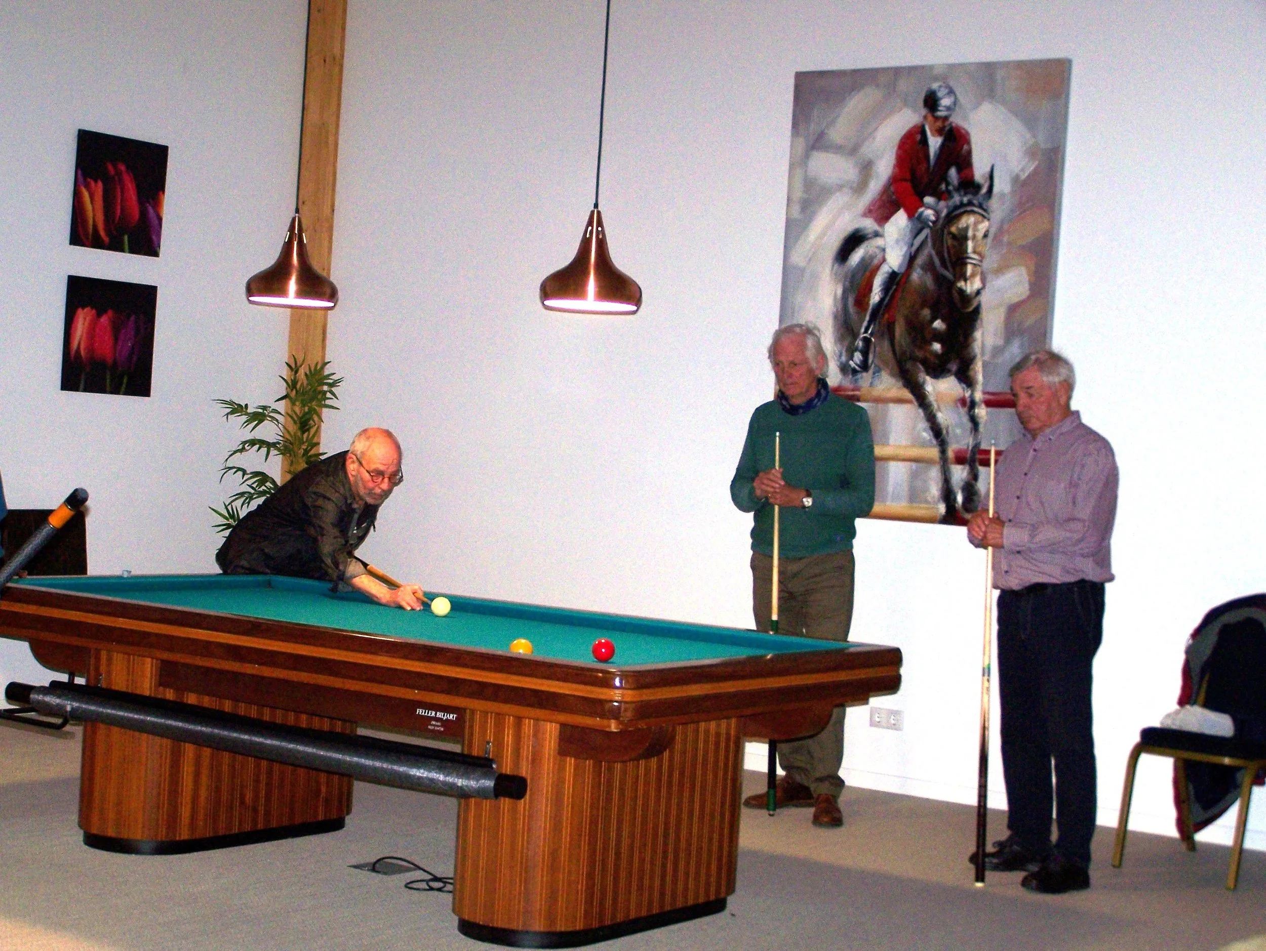 Three elderly men in a room with artwork, one playing billiards, two holding cue sticks, and a large painting of a jockey on a horse hanging on the wall.