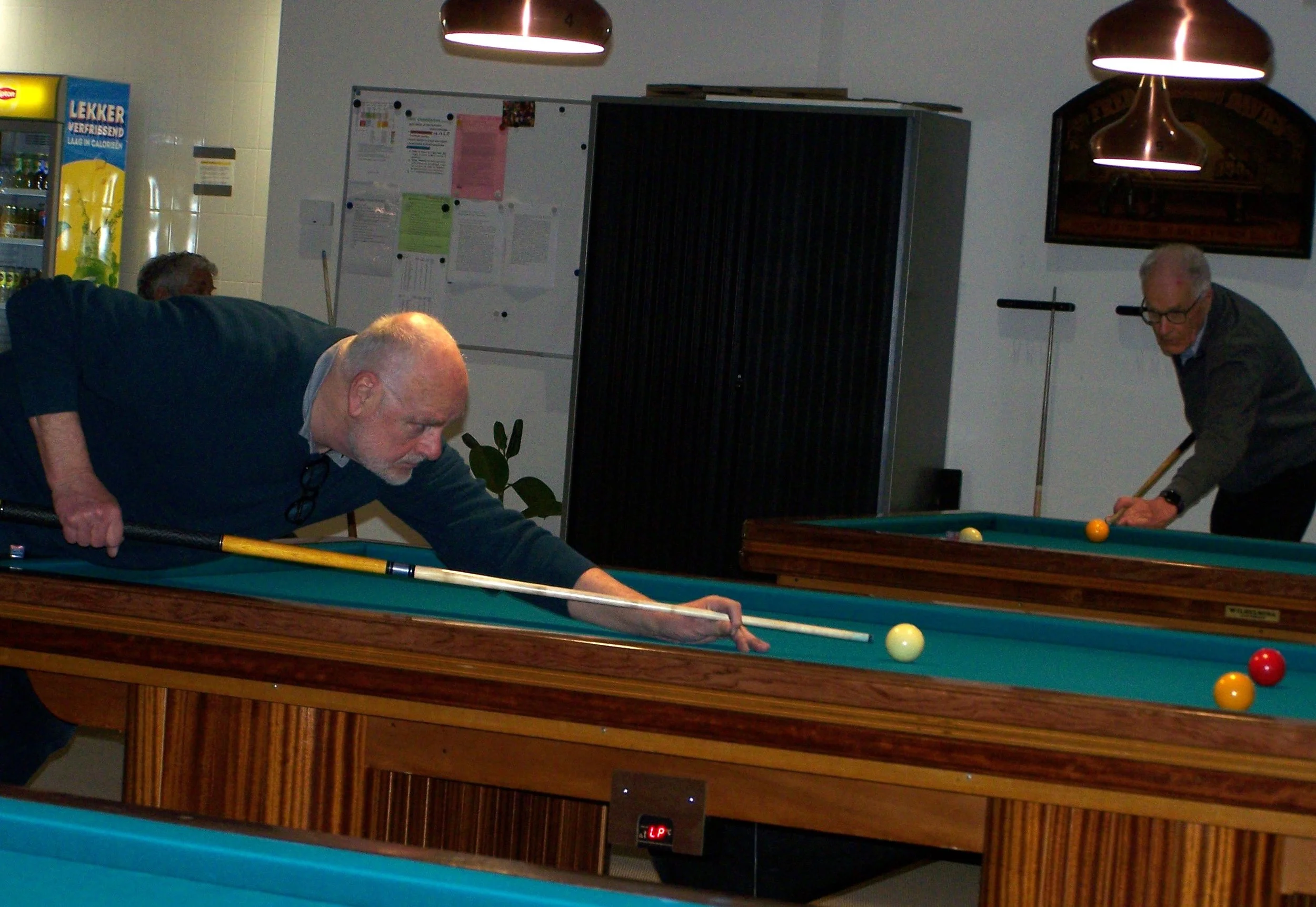 Two older men playing pool, one leaning over the table aiming the cue stick, the other preparing to take a shot, in an indoor recreational room.