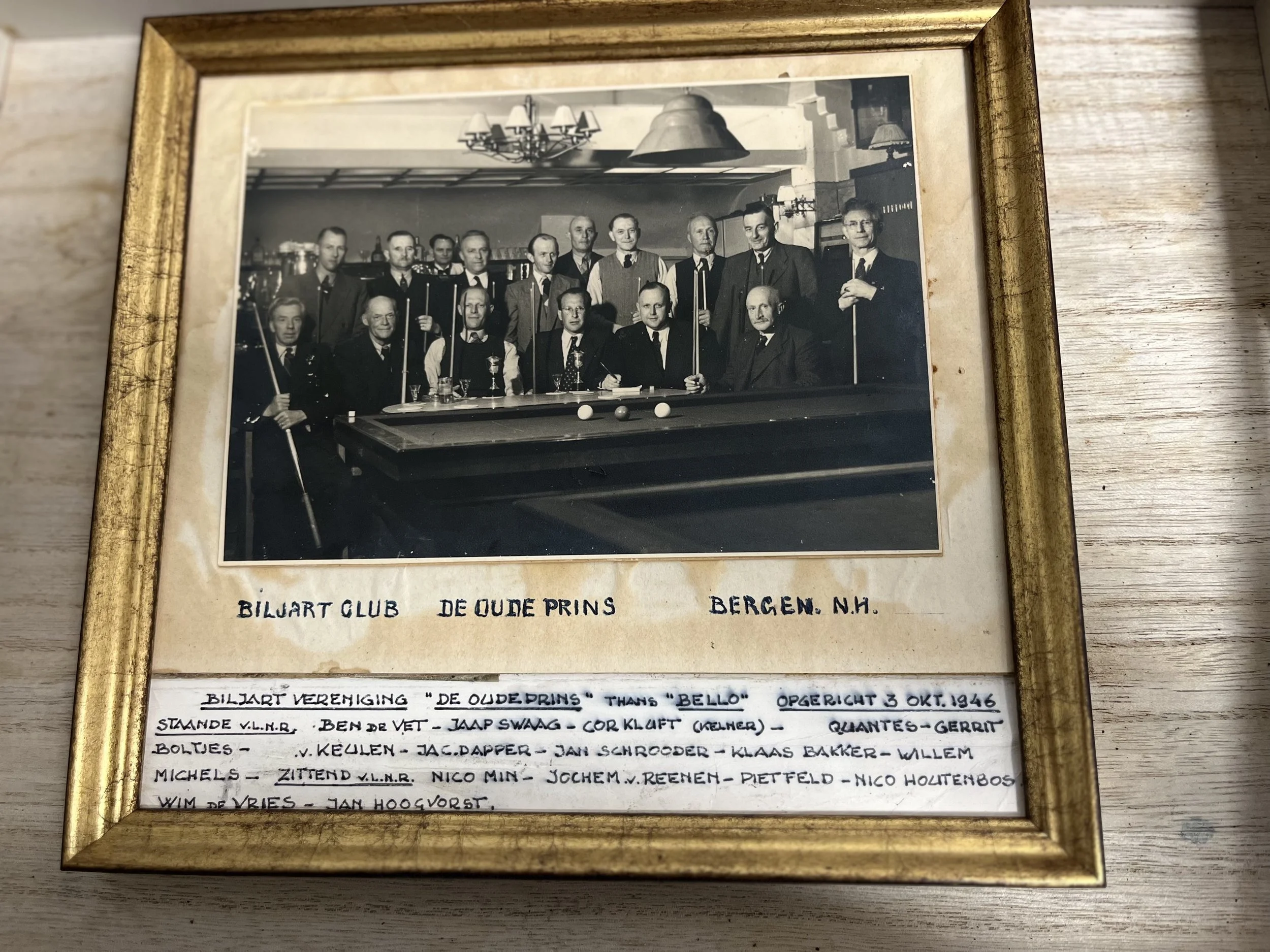 A framed black and white photograph of a group of men gathered around a billiards table, some holding pool cues, in a room with a chandelier and wall lights. Below the photo, handwritten text reads 'Biljart Club De Oude Prins Bergen N.H.' and additional notes about the club and members.