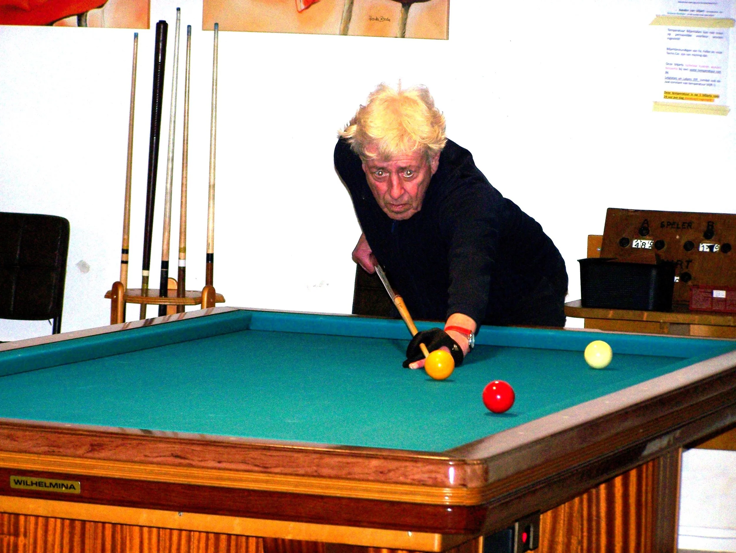 An elderly man with blond, tousled hair playing billiards on a green felt table, aiming at the yellow ball with a cue stick, wearing a black shirt and fingerless gloves.