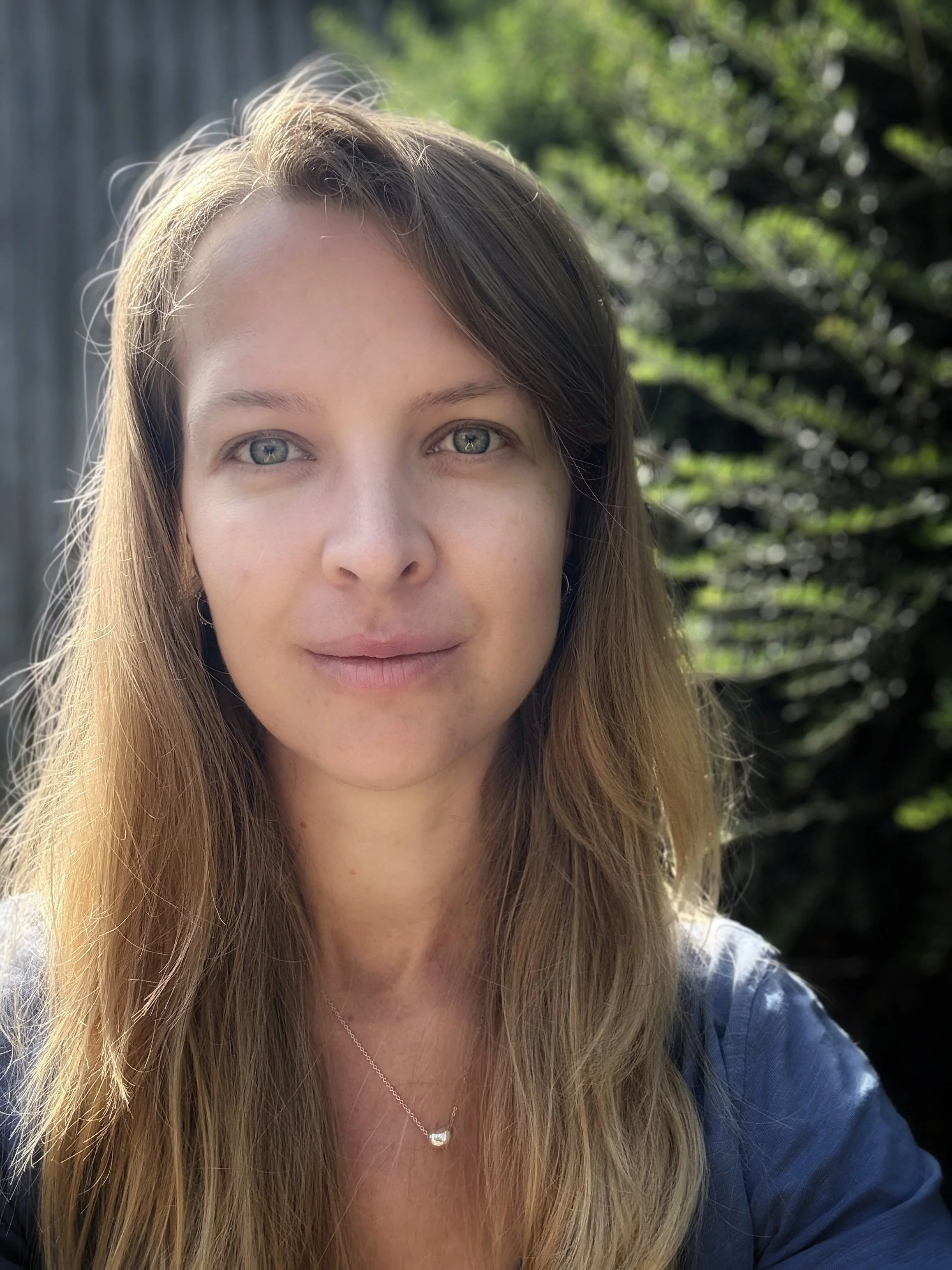A woman with long light brown hair and blue eyes taking a selfie outdoors on a sunny day with green foliage in the background.
