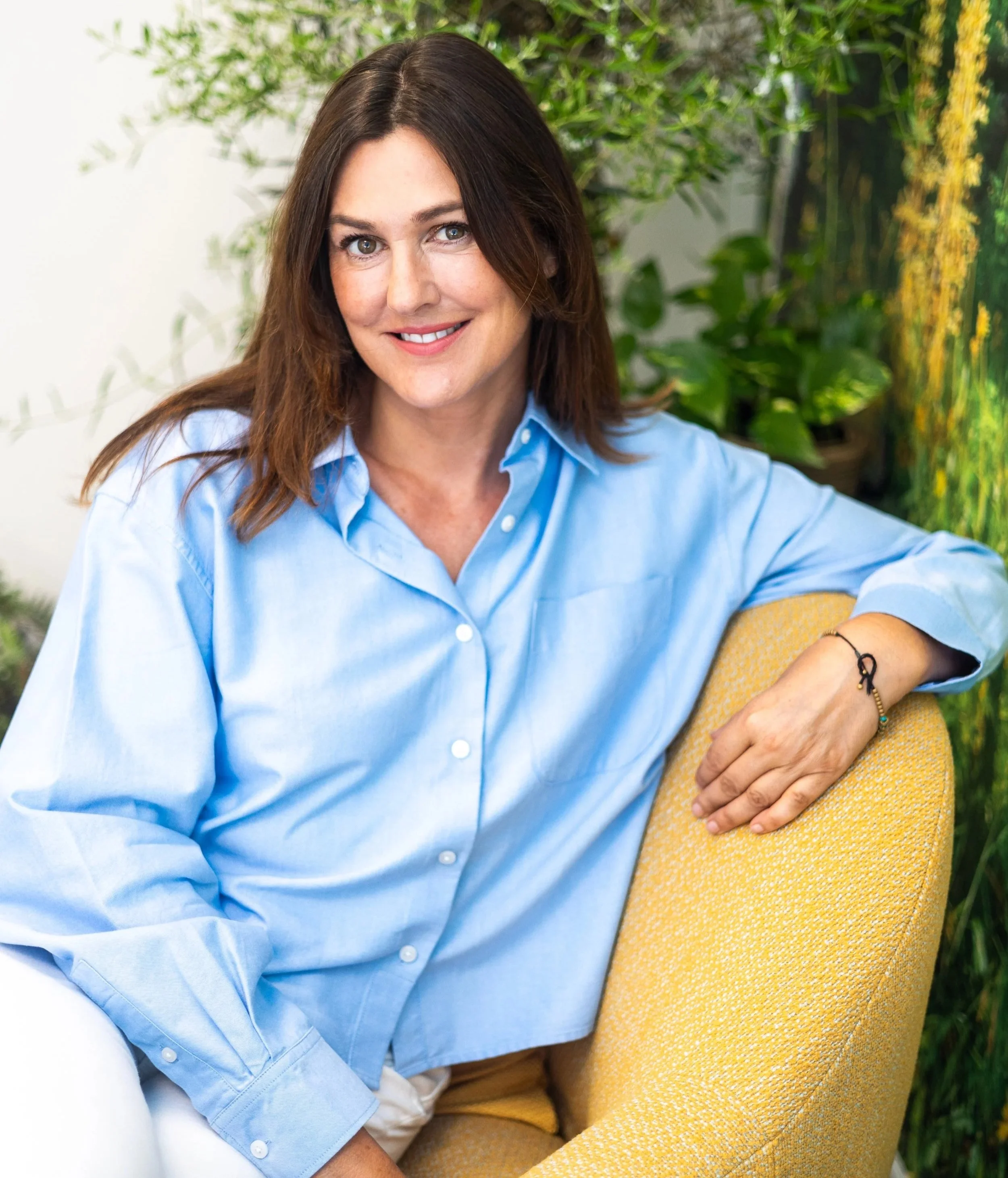 A woman with brown hair and blue eyes sitting on a yellow chair in a room with green plants and a colorful background, wearing a light blue button-up shirt and smiling at the camera.