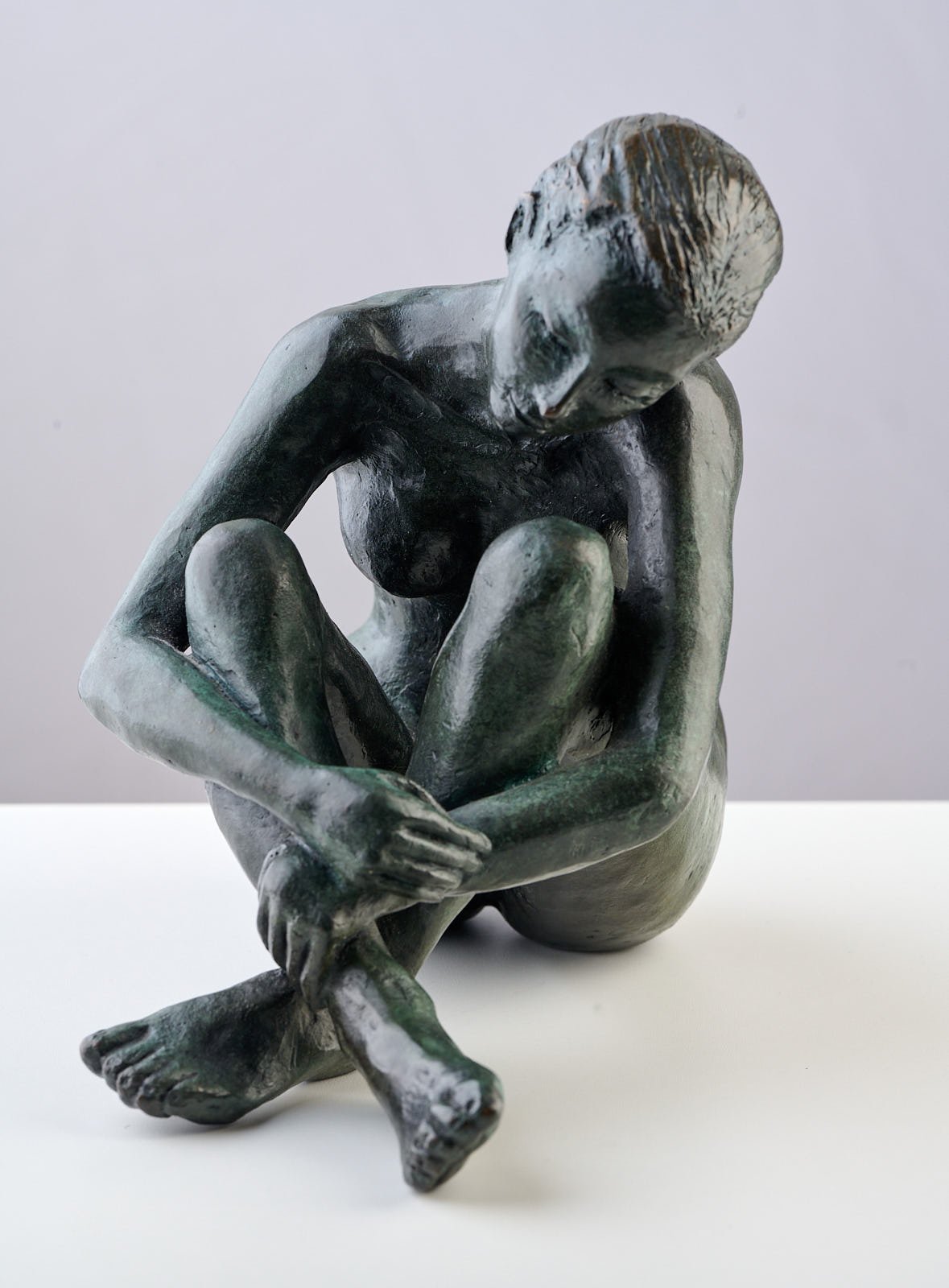Bronze sculpture of a seated woman with her head bowed and arms wrapped around her legs, on a white surface with a plain background.