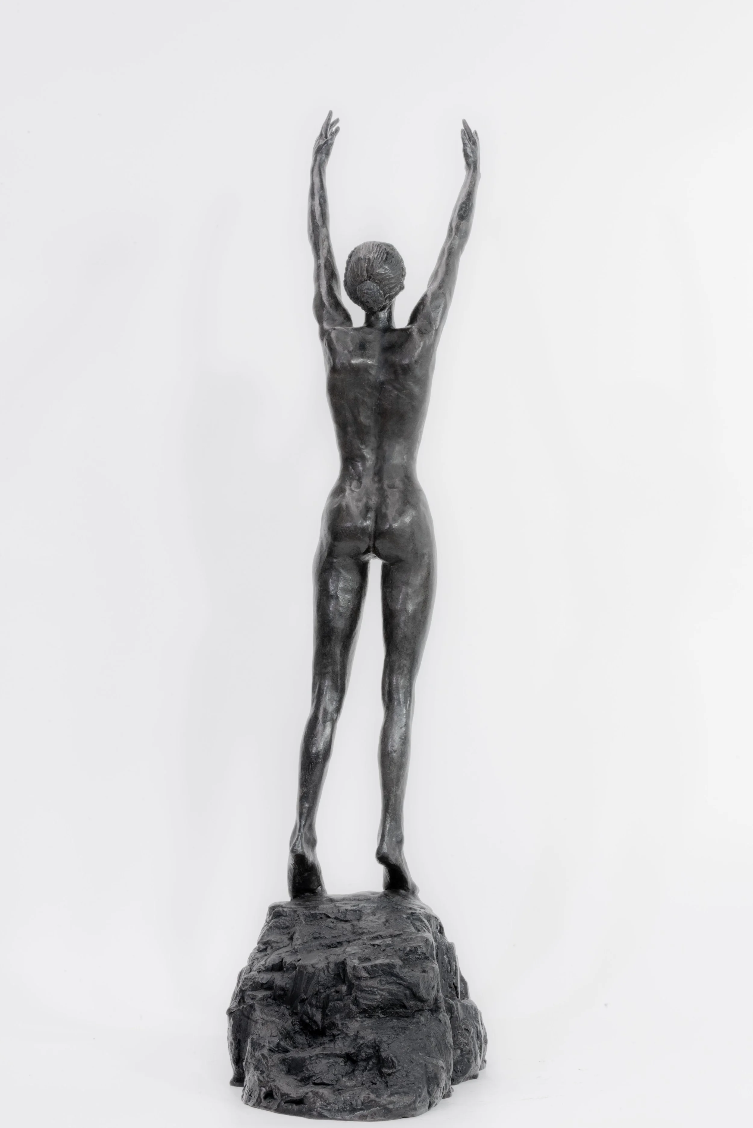 Bronze statue of a nude woman with arms raised above her head, standing on a rocky base against a plain white background.