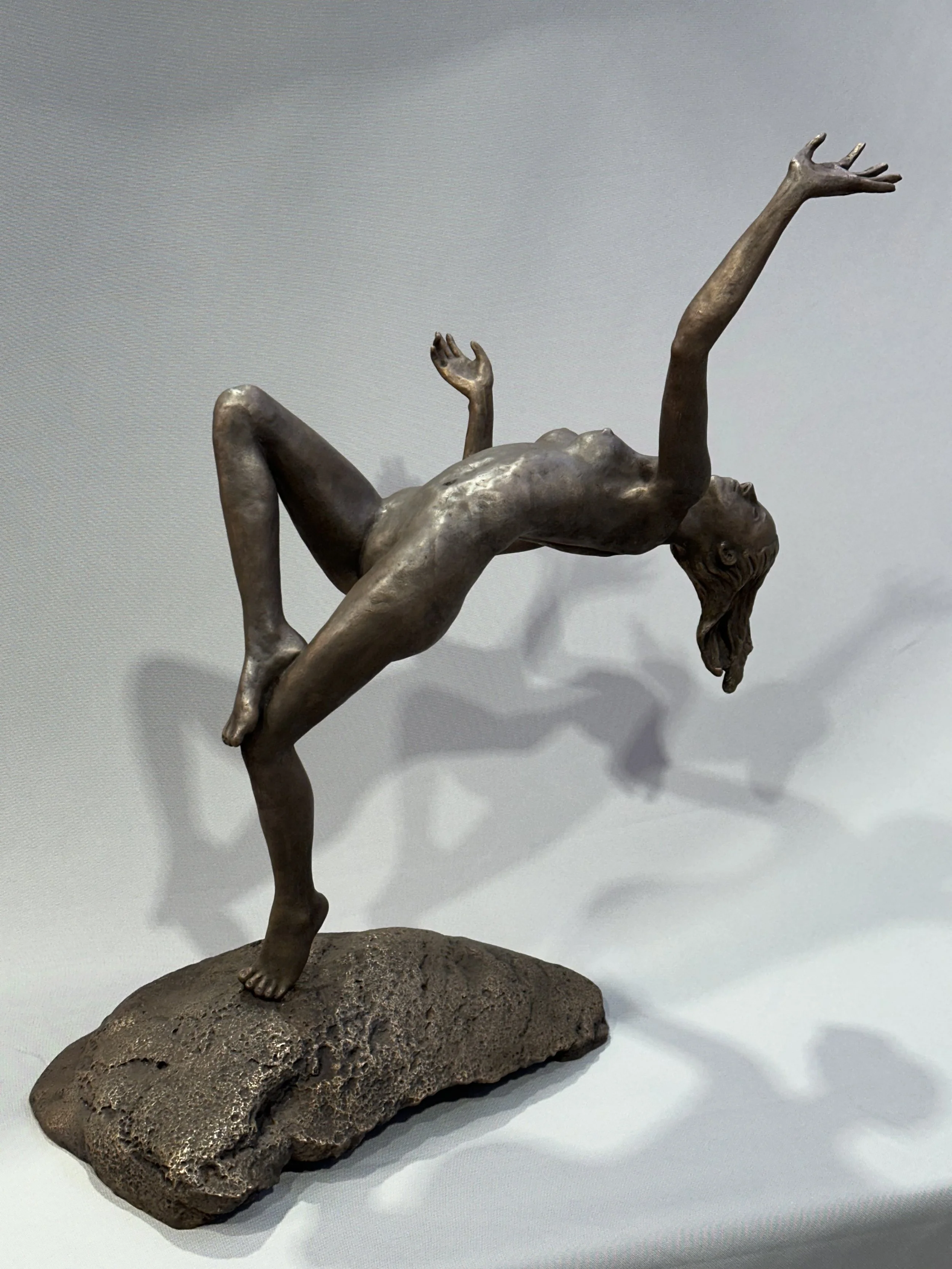 Bronze sculpture of a woman balancing on one hand with her body arched back and head tilted, standing on a textured rock base.