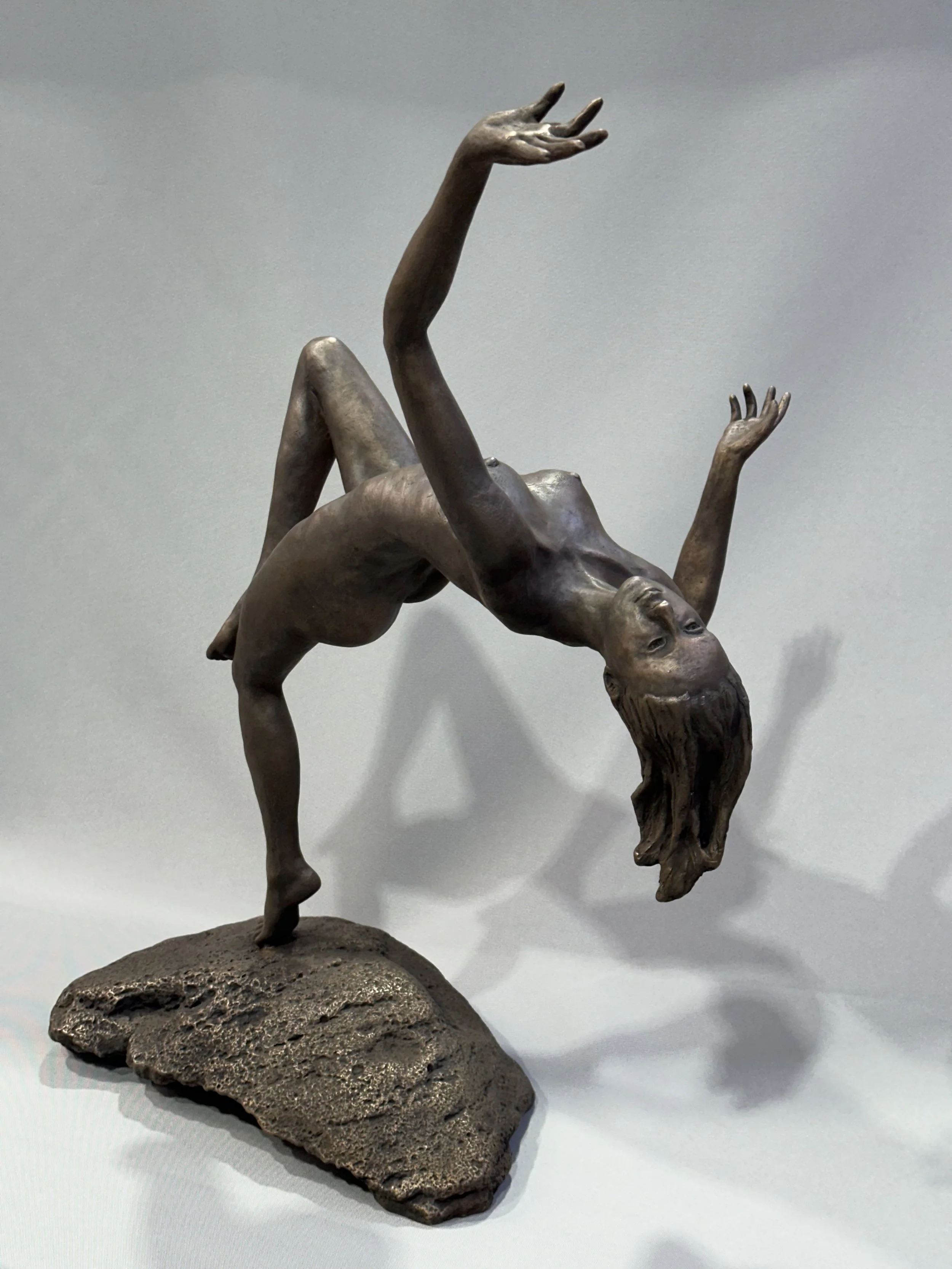 Bronze sculpture of a woman performing a backbend on a rock base, with her hair hanging downward and arms raised, casting a shadow on a plain background.