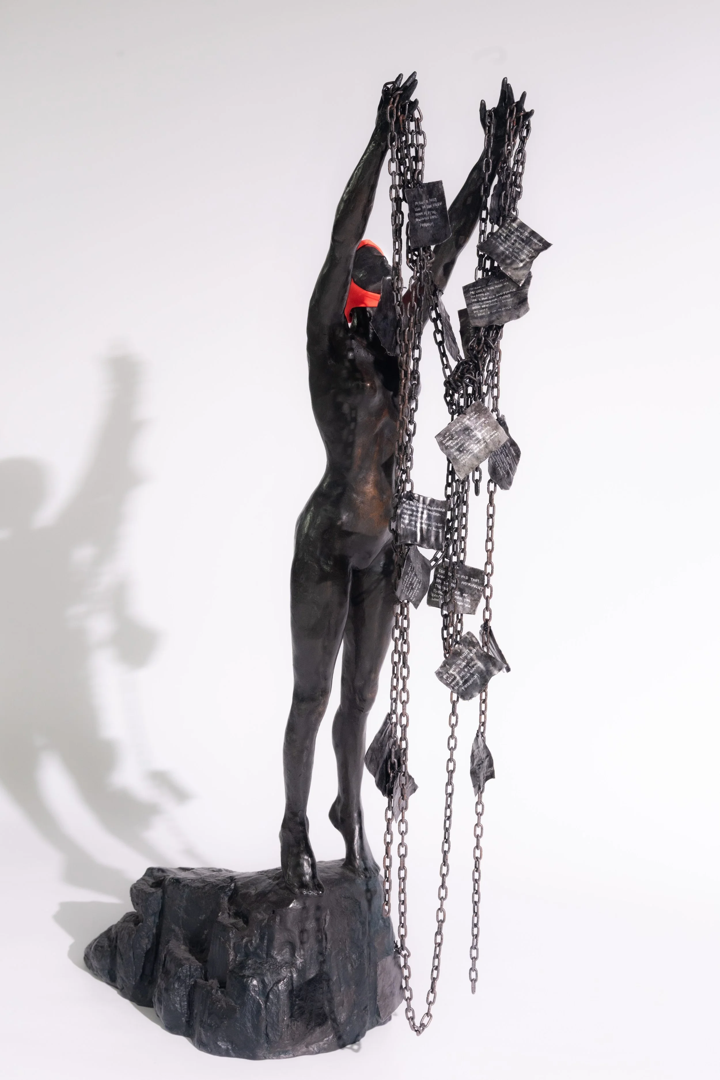 A dark sculpture of a woman with a red mask standing on a rocky base, holding chains with metal plates hanging from them.