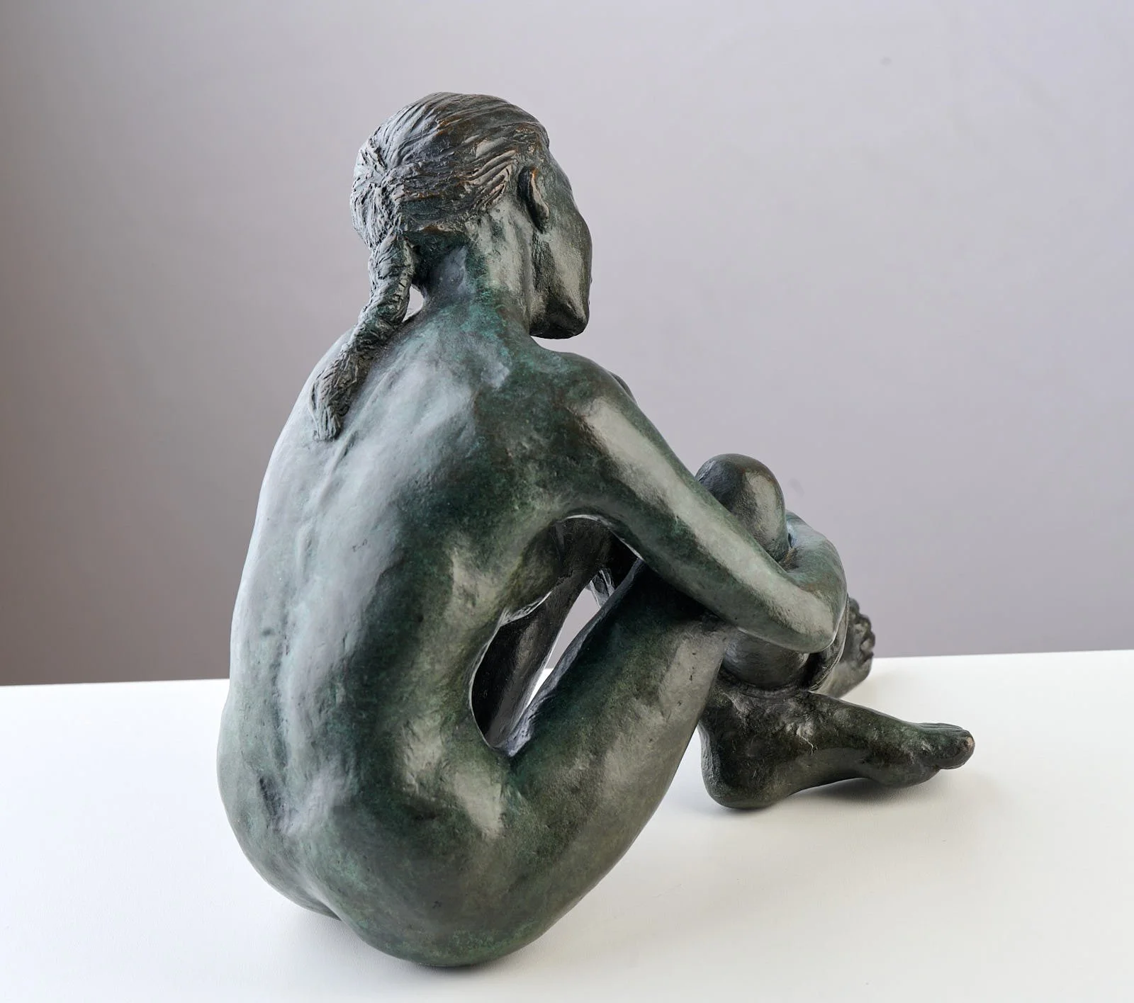 Bronze sculpture of a seated woman with her arms wrapped around her legs, sitting on a white surface against a gray background.