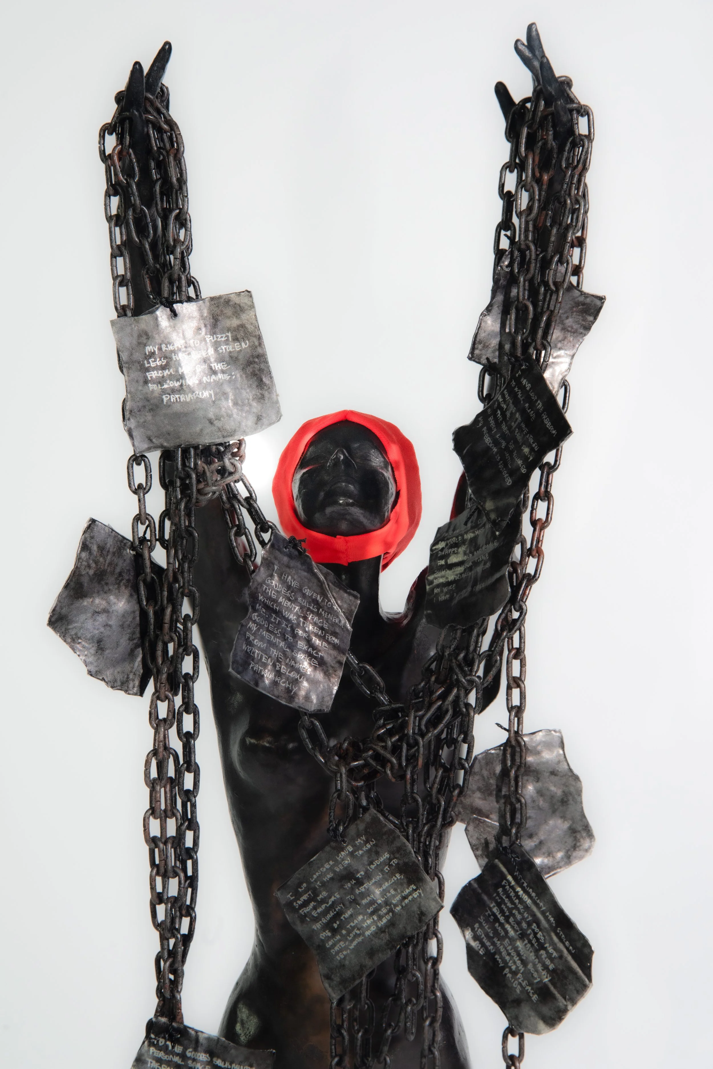 Sculpture of a black human figure with a red hood, wrapped in black chains with black metal plaques containing text, set against a white background.