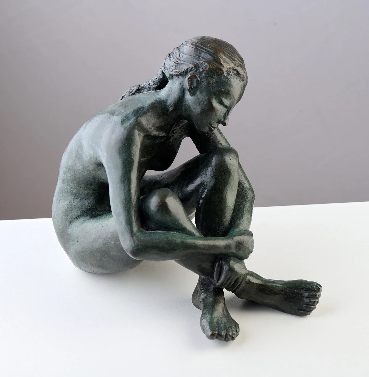 Bronze sculpture of a woman sitting with her arms wrapped around her bent legs, head bowed, on a white surface against a gray background.