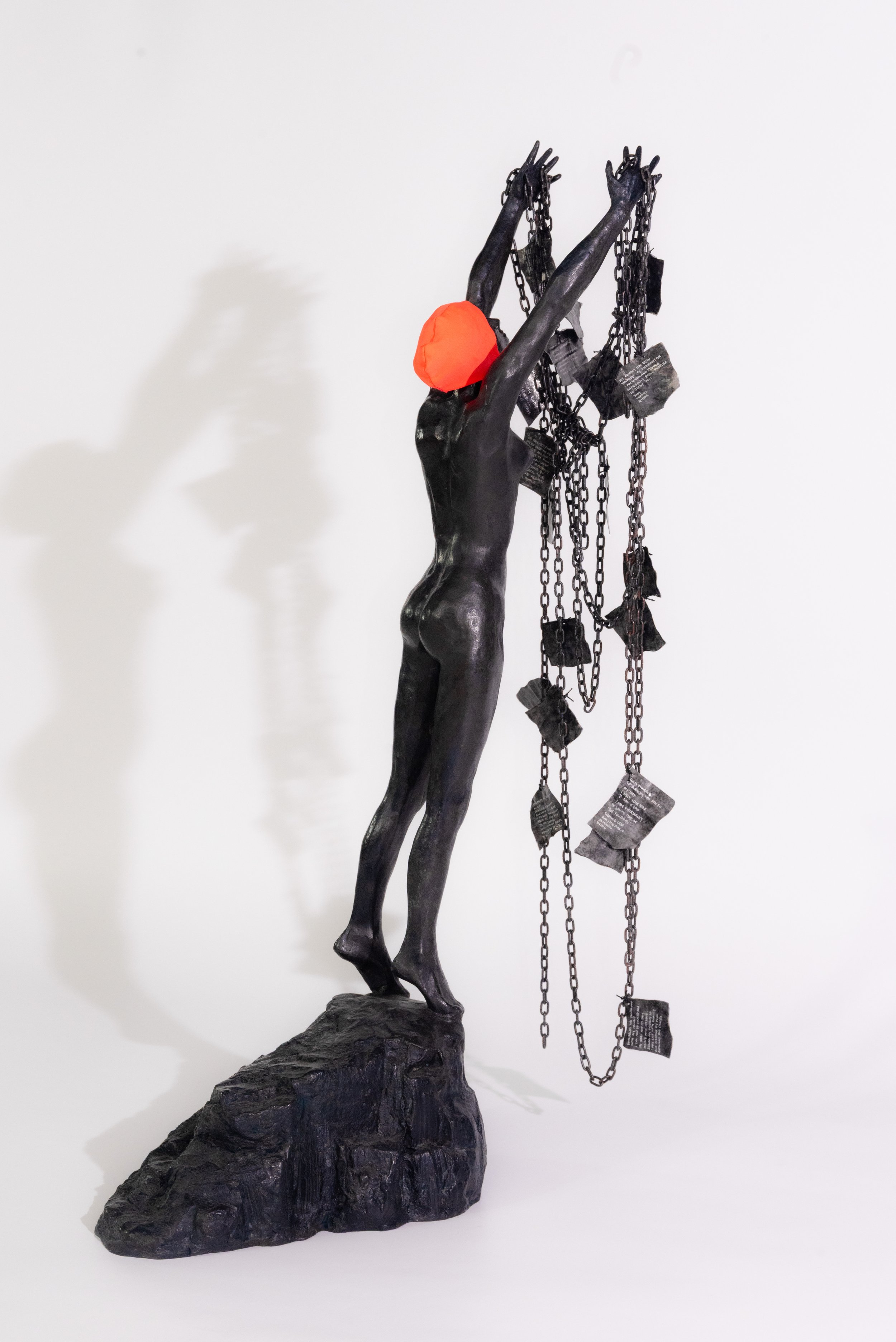 Sculpture of a black female figure with a red head covering, standing on a rocky base, reaching up to hold hanging chains and paper-like objects.