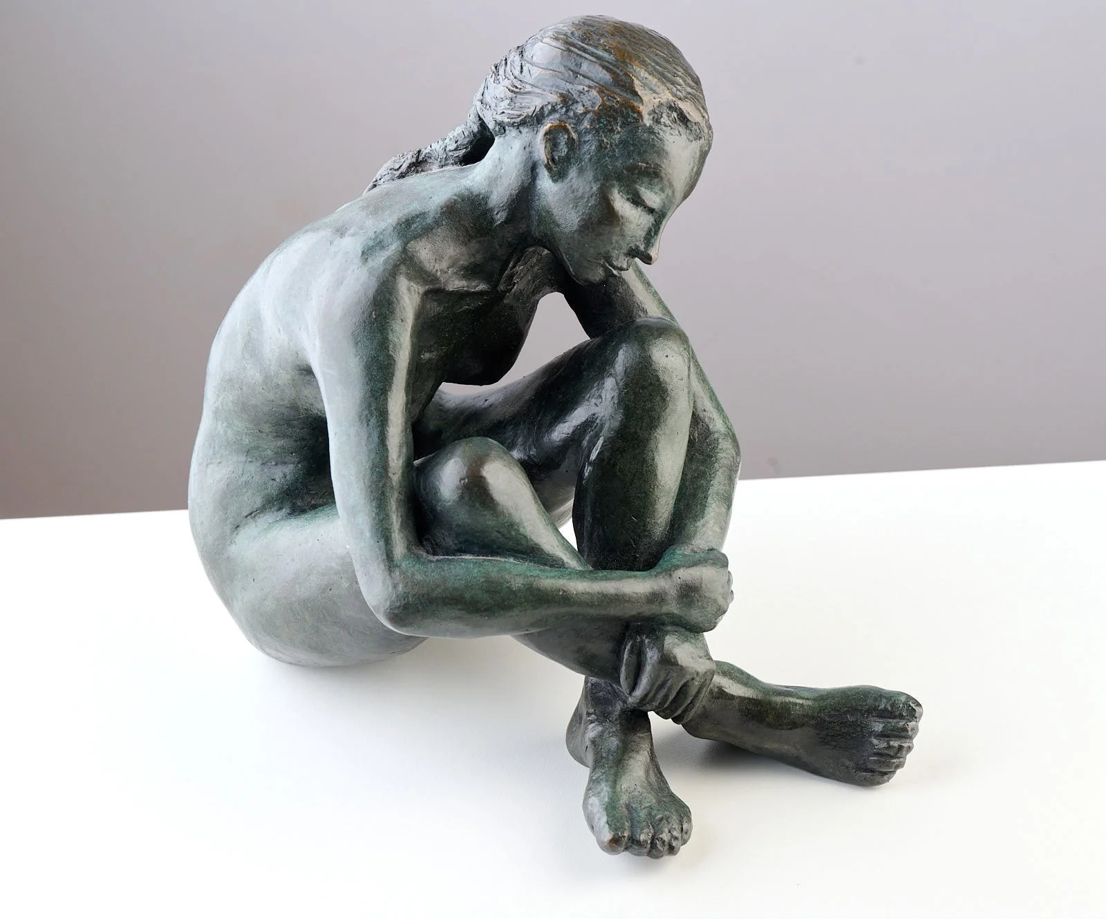 Bronze sculpture of a woman sitting with one leg crossed over the other, resting her head on her knee, with a contemplative expression.