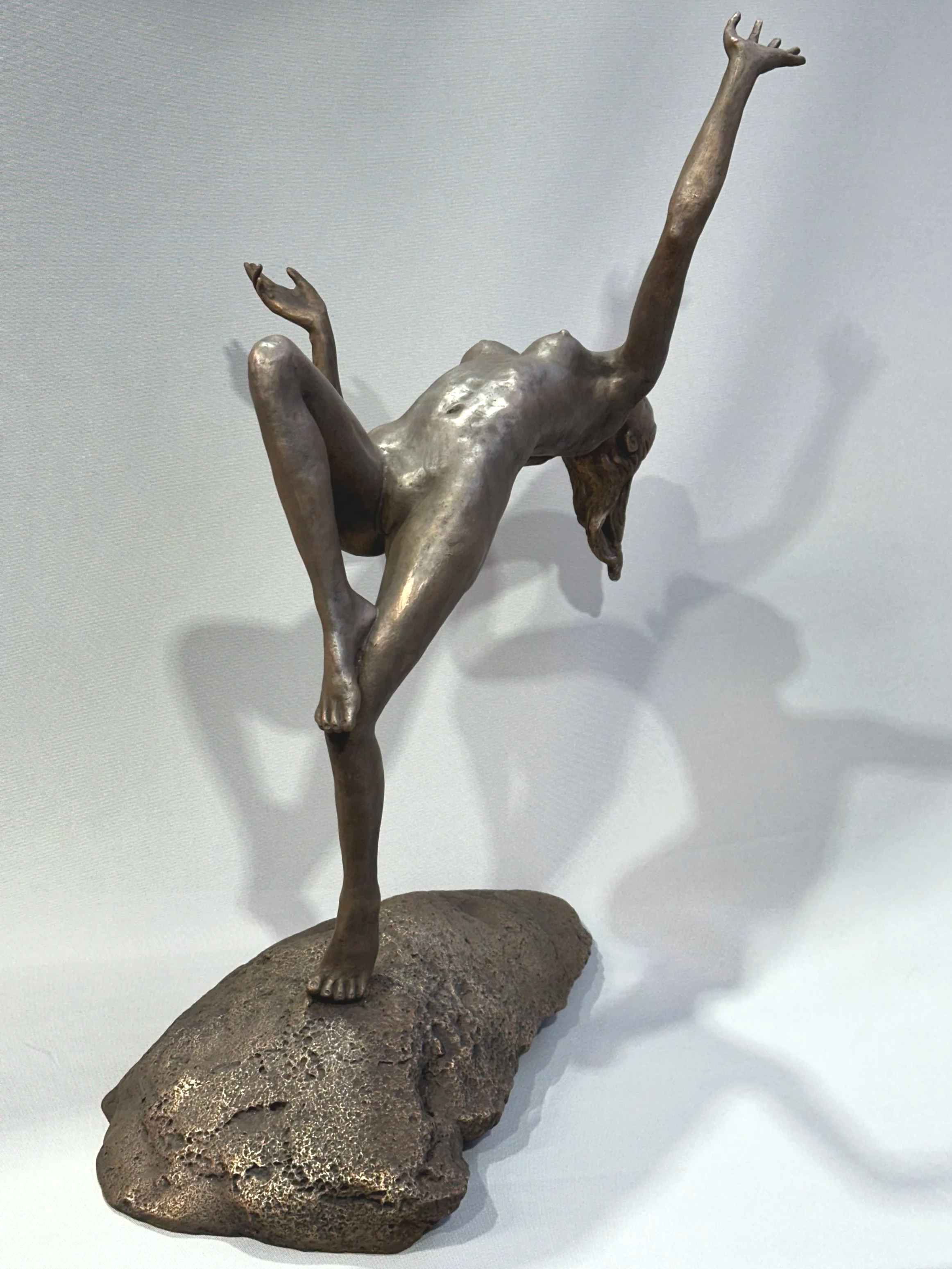 A sculpture of a woman balancing on one leg with the other leg bent and raised, one arm reaching upward and the other bent to the side, mounted on a textured rock base.