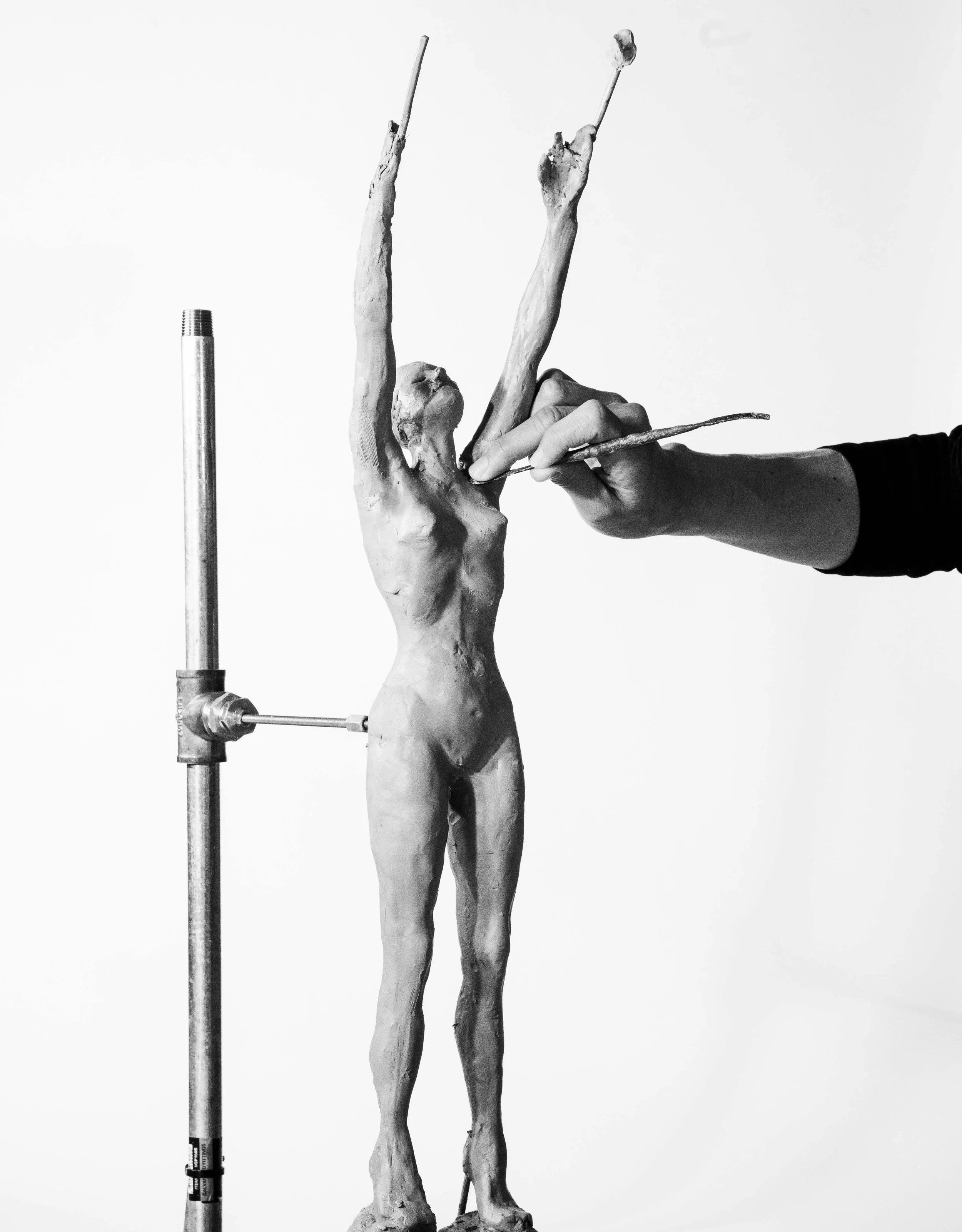 Black and white photo of an artist sculpting a human figure statue using clay and tools, with a support stand to hold the sculpture upright.