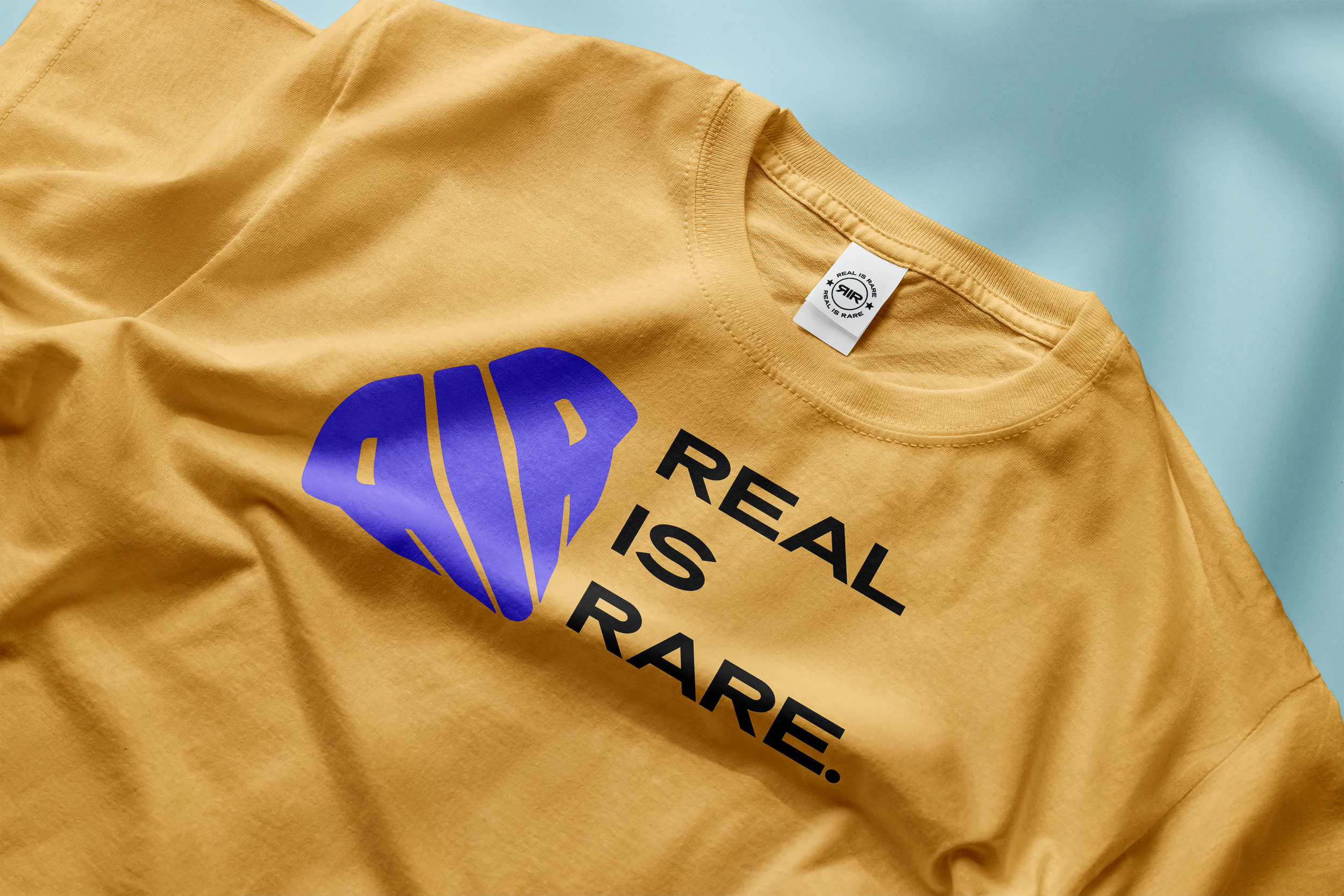 Geel T-shirt met een blauw superhelden-stijl diamantlogo en de tekst "REAL IS RARE", plat liggend op een lichtblauwe ondergrond.