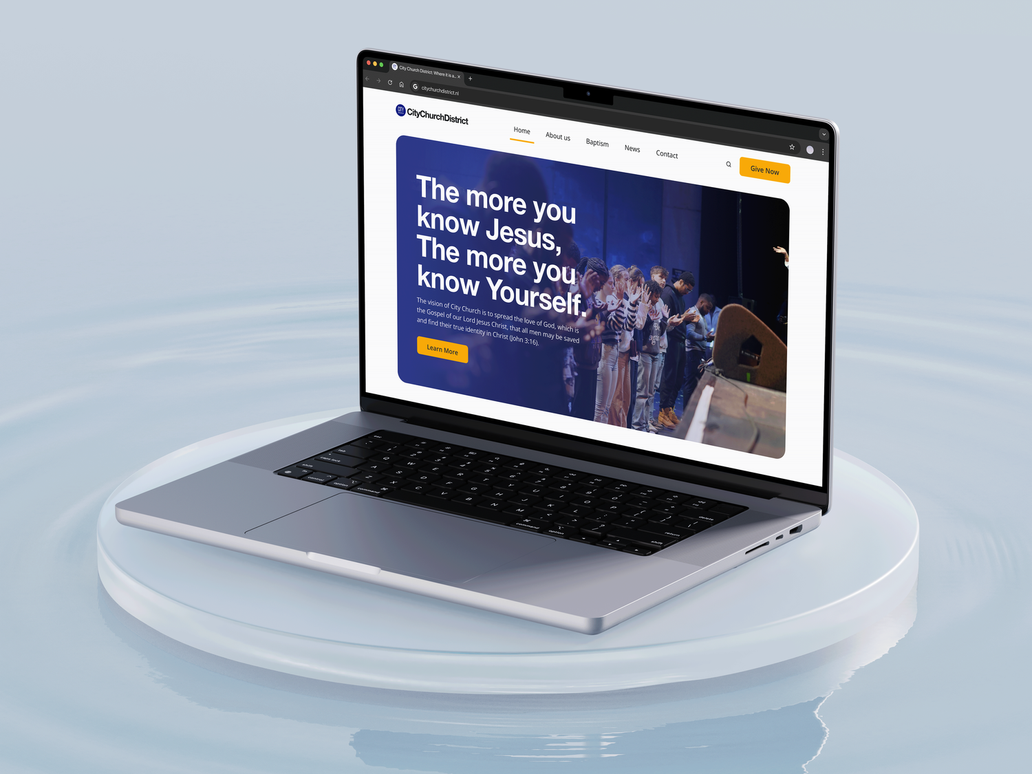 Zilveren MacBook-laptop op een wit rond platform, met daarop een kerkwebsite met een groep kinderen en de tekst: 'Hoe meer je Jezus kent, hoe meer je jezelf kent.'