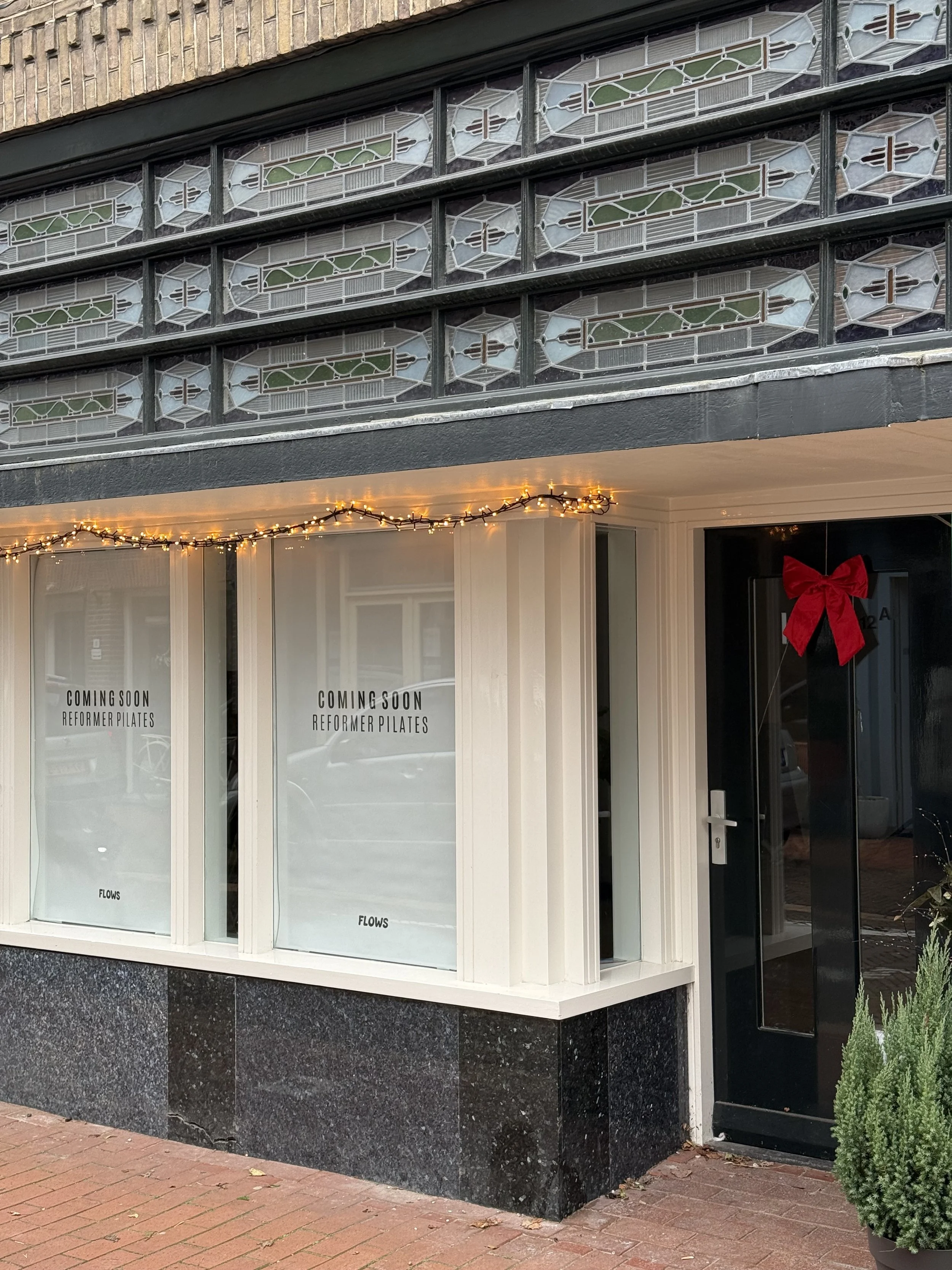 Voorgevel van een pilates studio in bussum met een raam waarop staat 'COMING SOON REFORMER PILATES', versierd met kerstverlichting en een rode strik aan de deur, naast een kleine groene struik.