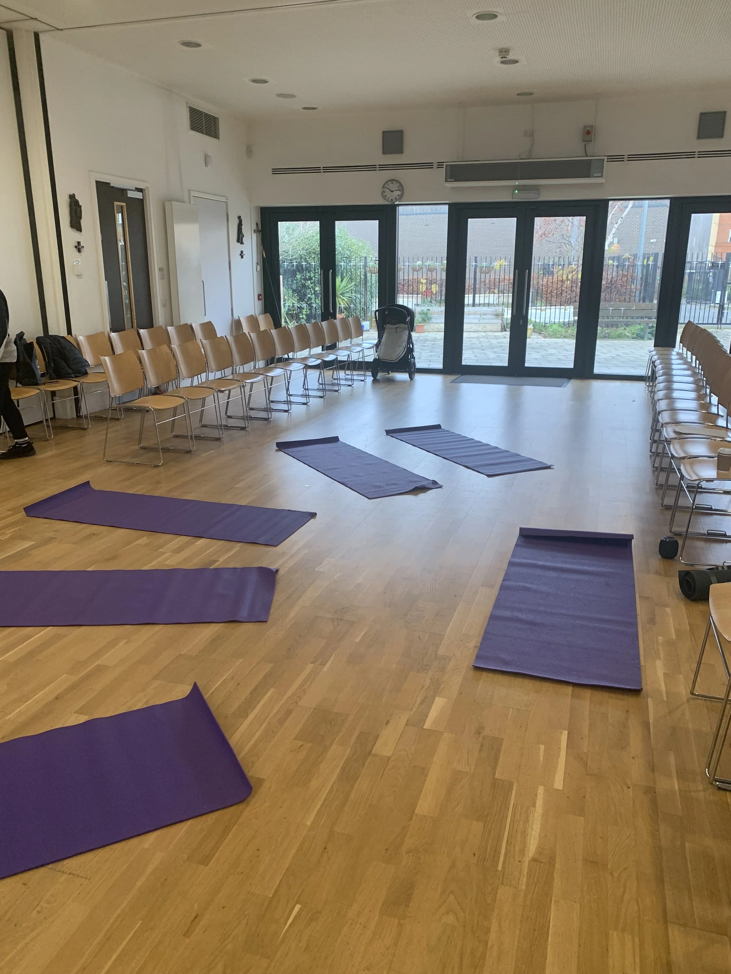 A spacious room set up for a yoga class with purple mats arranged on a wooden floor, surrounded by chairs along the walls, large glass doors opening to an outdoor garden area, and a stroller near the doors.