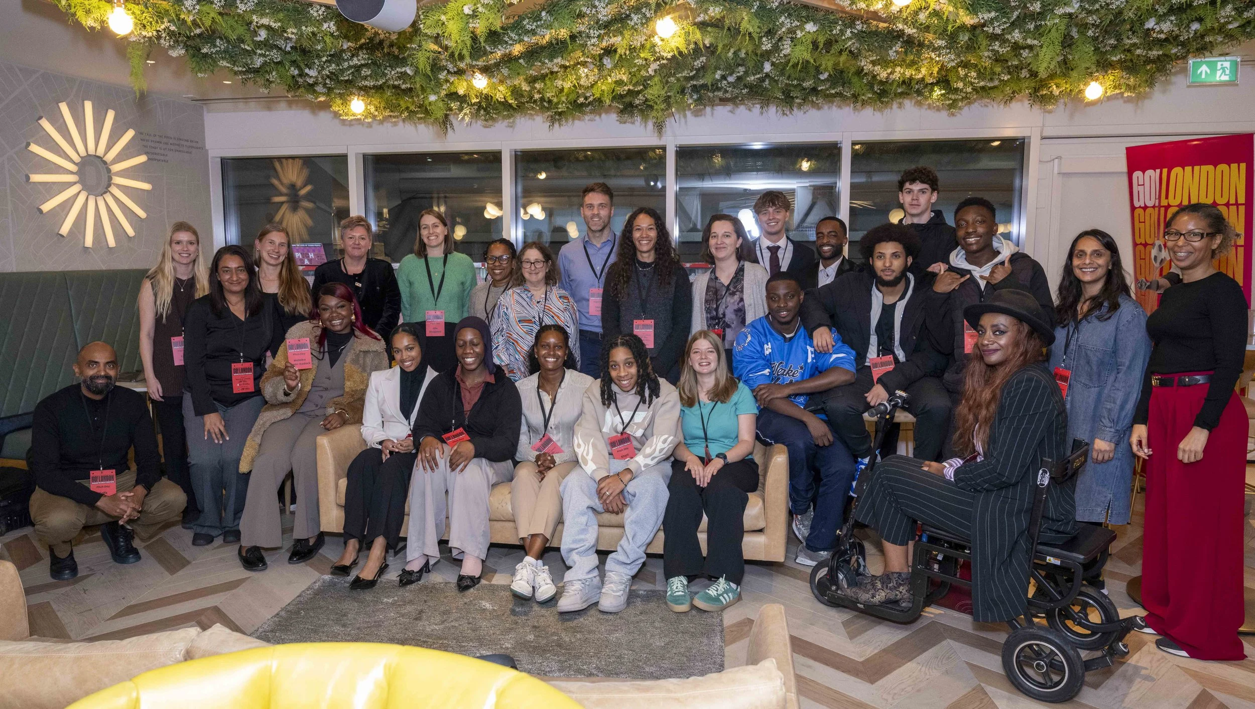 Group photo of the Go! London team, including young entrepreneurs, the judging panel and Go! London staff. There is festive decoration above and a red banner on the right that says "GO! LONDON." Everyone is smiling and wearing name tags.
