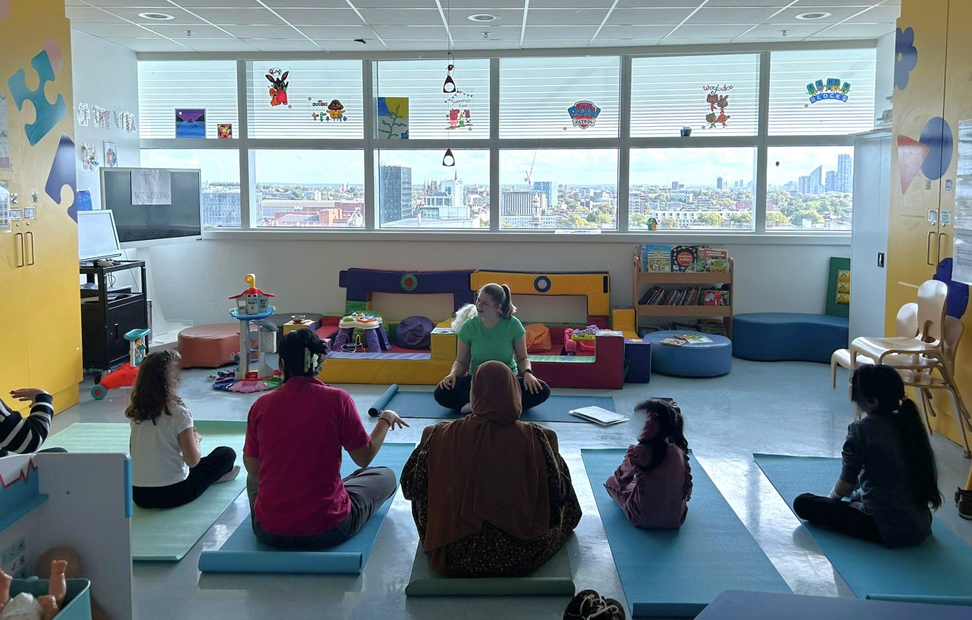 A group of adults and children participating in a children's yoga class in a brightly lit room with large windows and a city view.