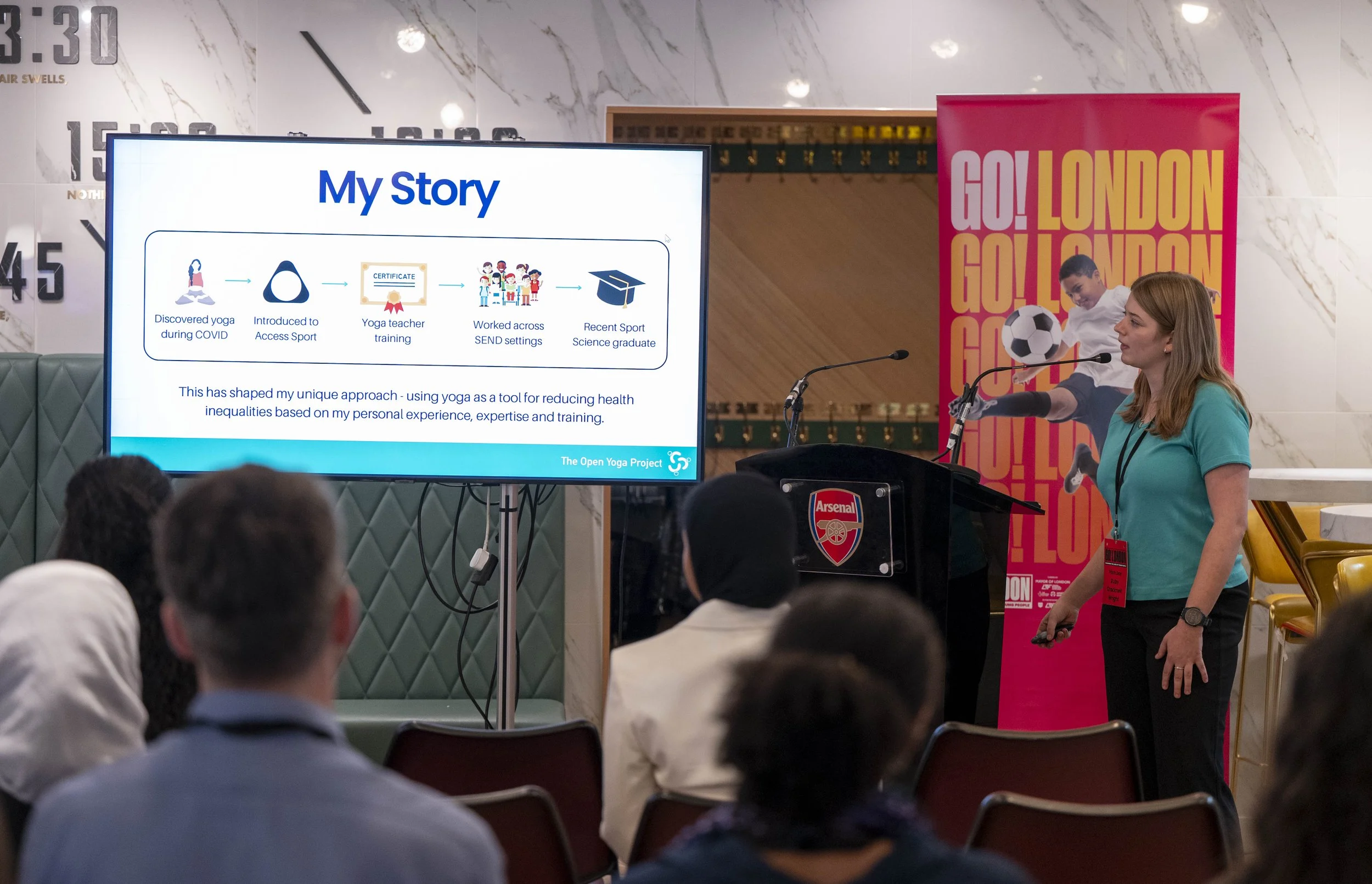 A woman in a turquoise shirt is giving a presentation to the Go! London Panel about her story on yoga and health, with a large screen displaying her story steps, and audience members seated and listening.