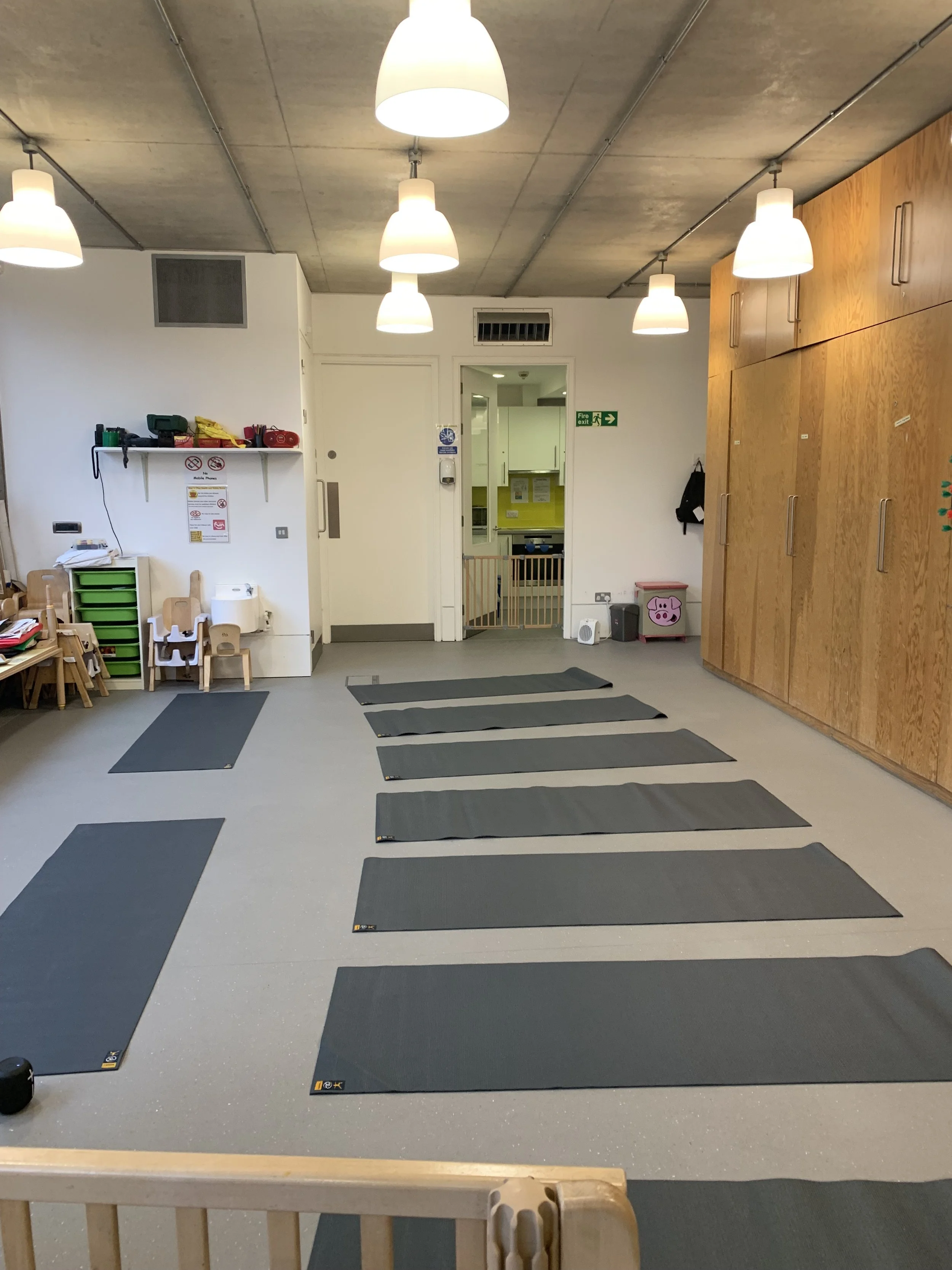A children's centre room set up for yoga with multiple black mats arranged on a grey floor, white walls, wooden cabinets on the right, and a ceiling with hanging white lights.