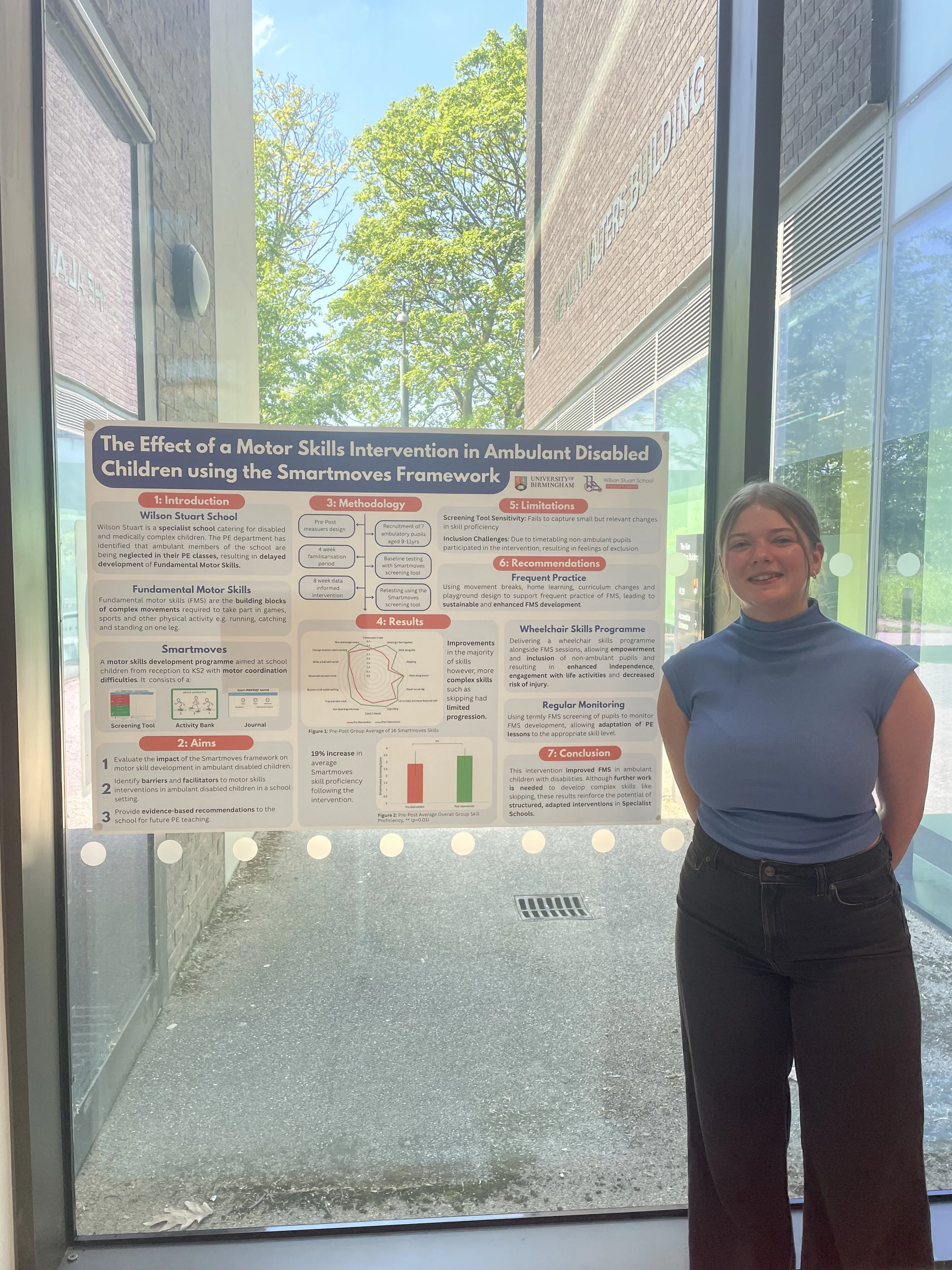 A woman in a blue shirt and black pants standing next to a research poster at a conference or presentation event, with trees and a building exterior visible through a glass window behind her.