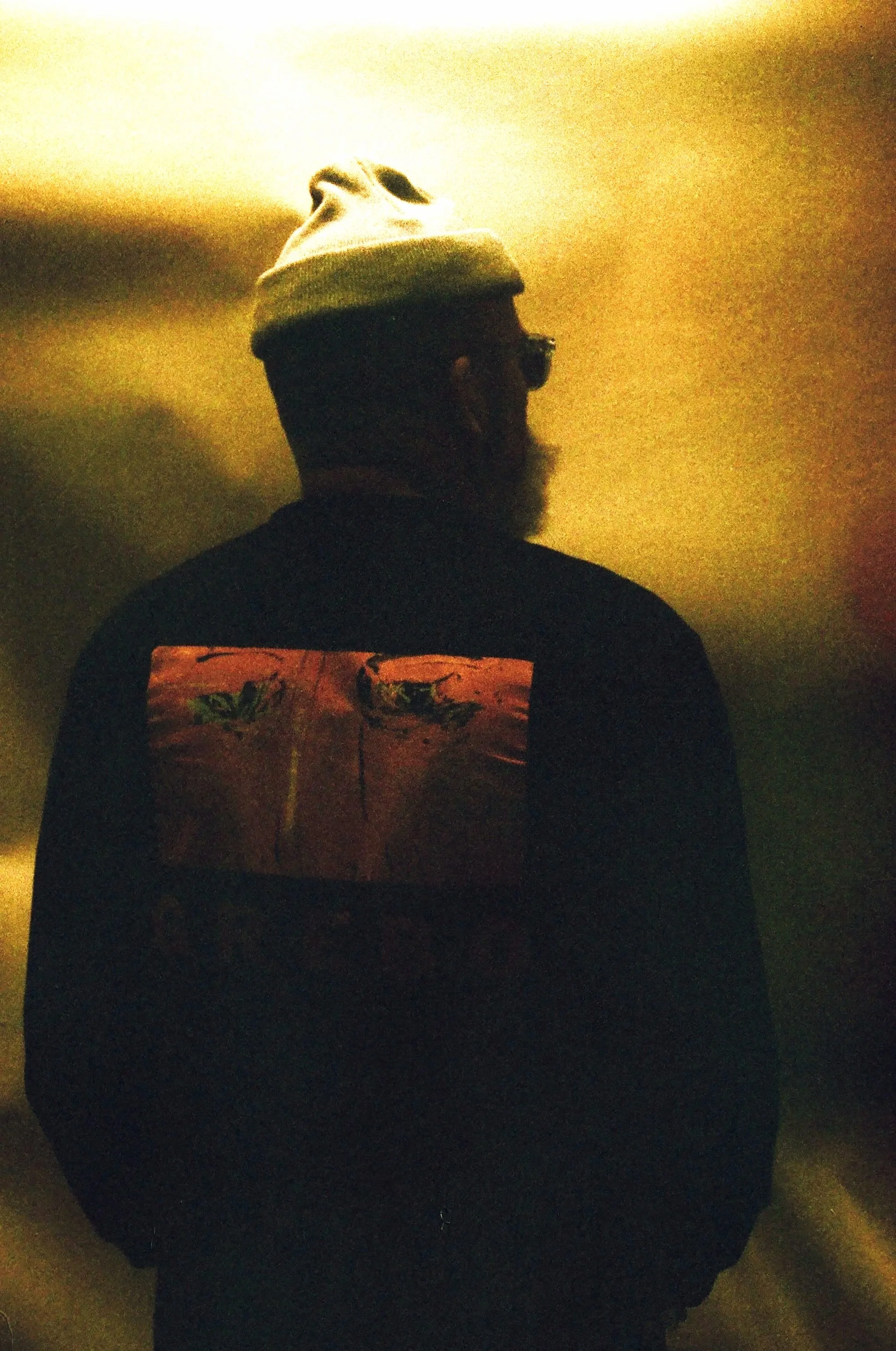 A person with a beard wearing a gray beanie and glasses, facing away, wearing a black jacket with a colorful design on the back, in a dimly lit environment.