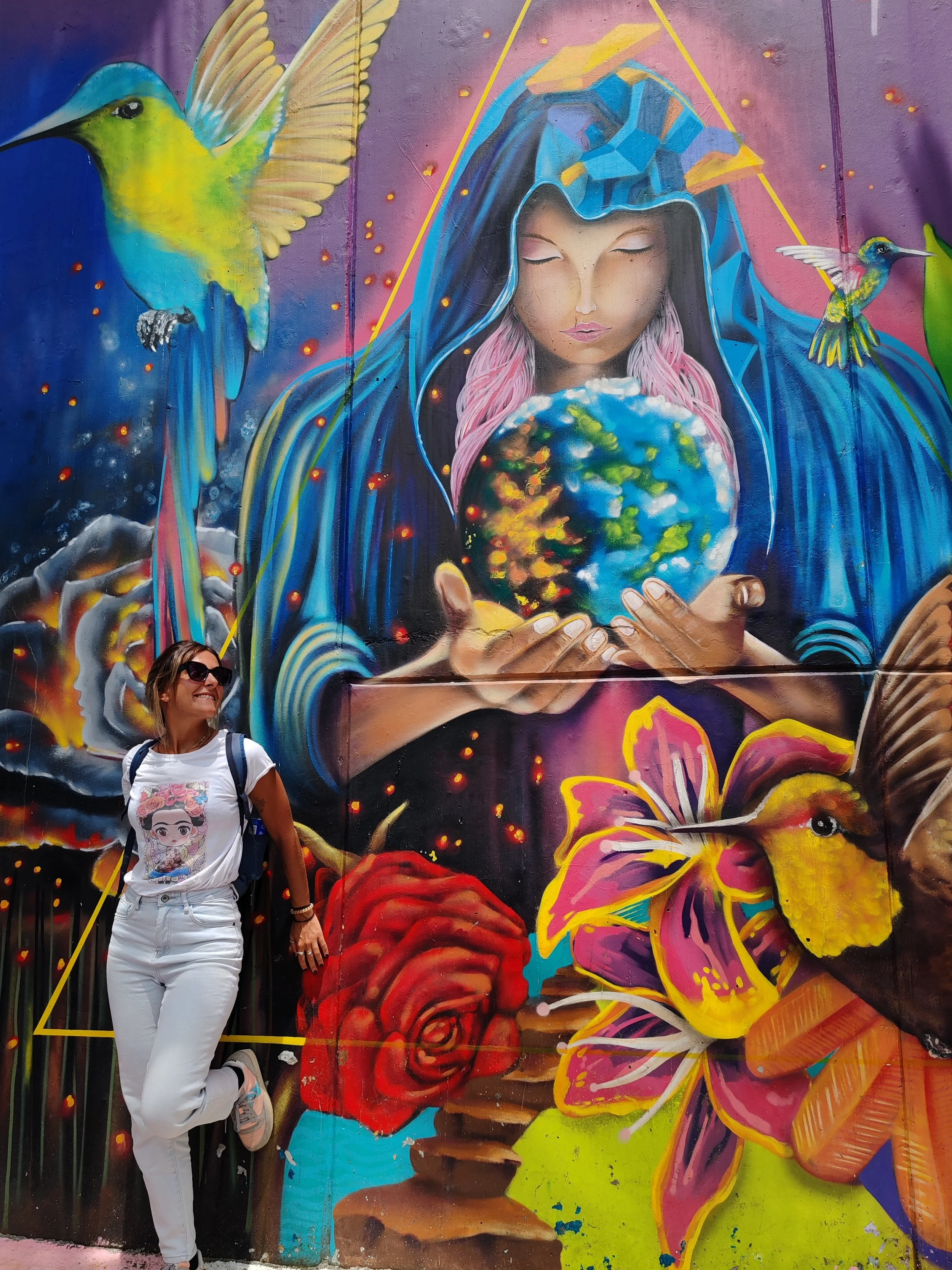 Una donna sorridente con occhiali da sole di fronte a un murale colorato in Colombia, a Medellin, che raffigura una figura femminile con un cappuccio blu, uccelli, fiori e una rappresentazione del pianeta Terra.