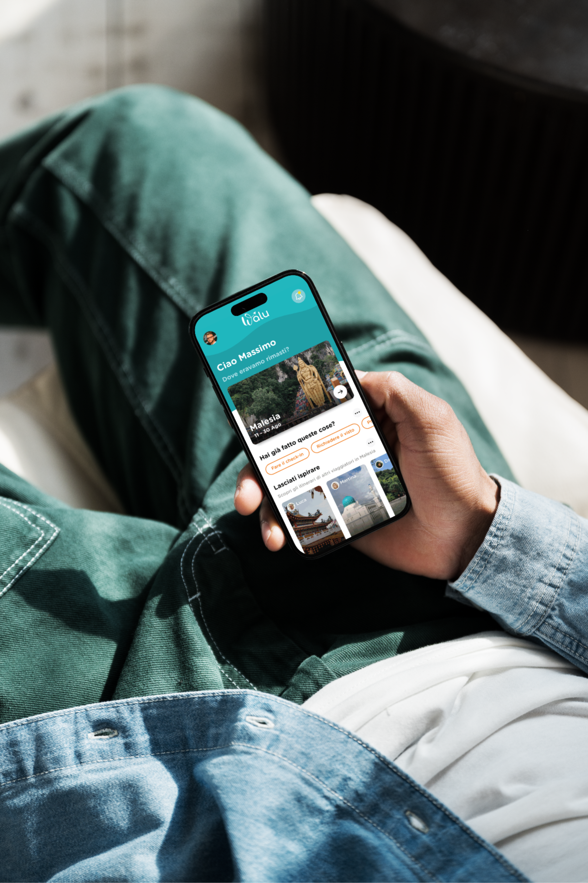 Persona seduta con pantaloni verdi e una camicia in denim chiaro, che tiene in mano uno smartphone che mostra l'app di viaggio Walu con la creazione di un itinerario per un viaggio in Malesia.