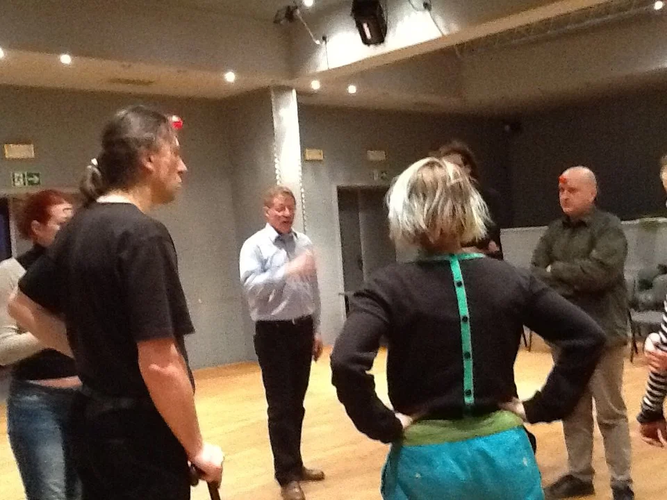 A group of people engaged in a conversation or discussion in an indoor setting, with some standing and one person gesturing with their hand.