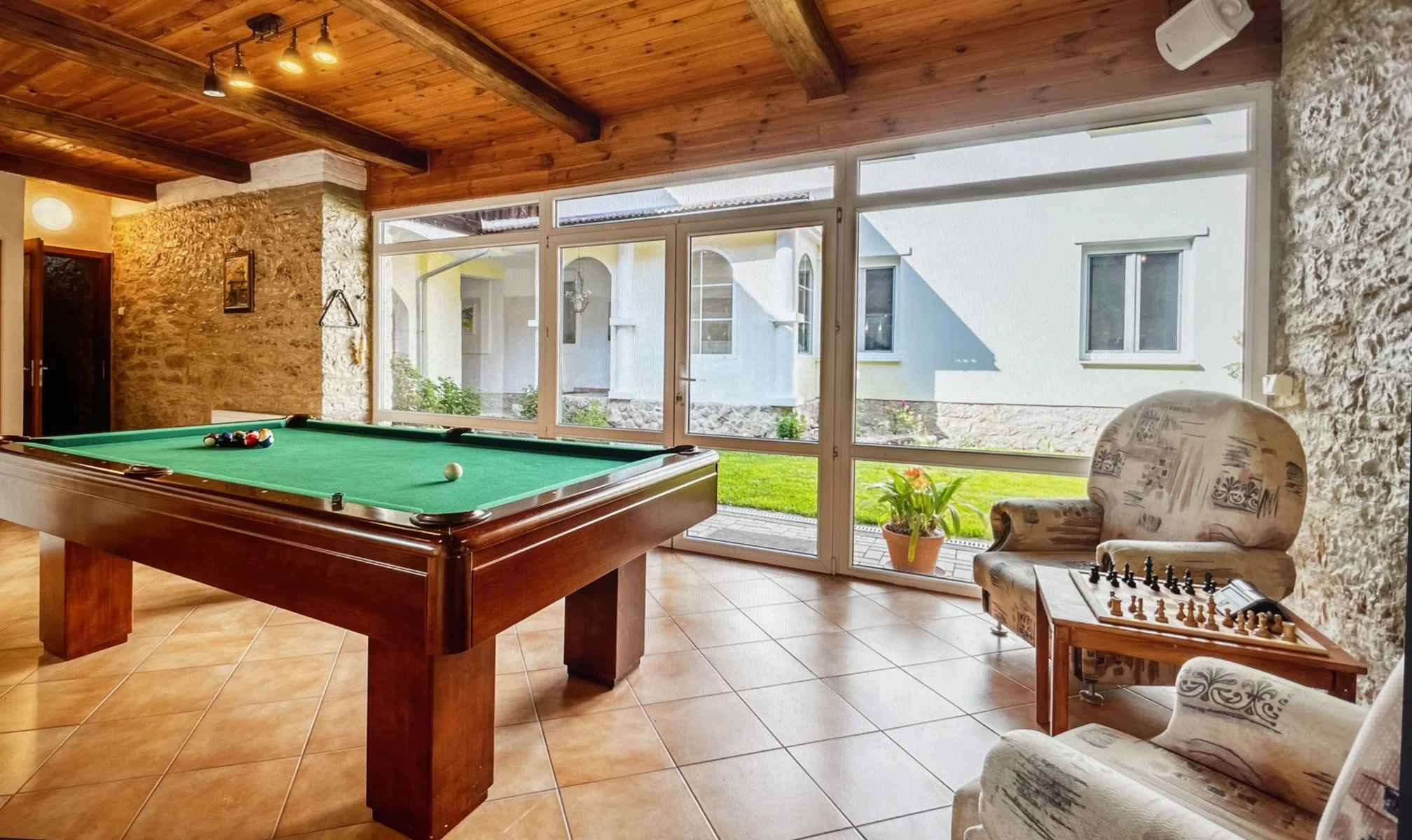 Indoor recreation room with a pool table, a chessboard on a sitting area, large windows revealing a green lawn outside, wood-paneled ceiling, and brick walls.