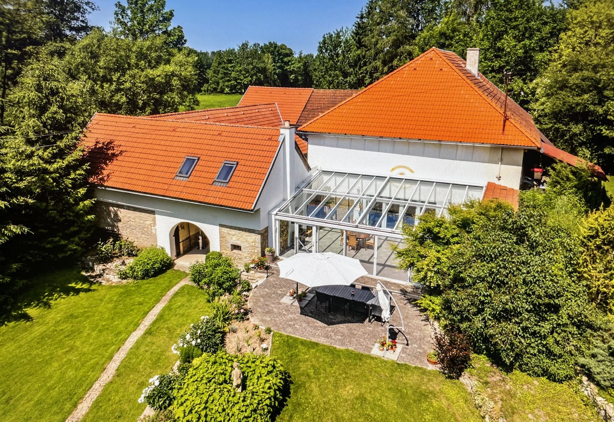 Aerial view of a house with red-tiled roof, white walls, and a glass conservatory, surrounded by a lush green garden with a patio, umbrella, and outdoor furniture in a rural setting.