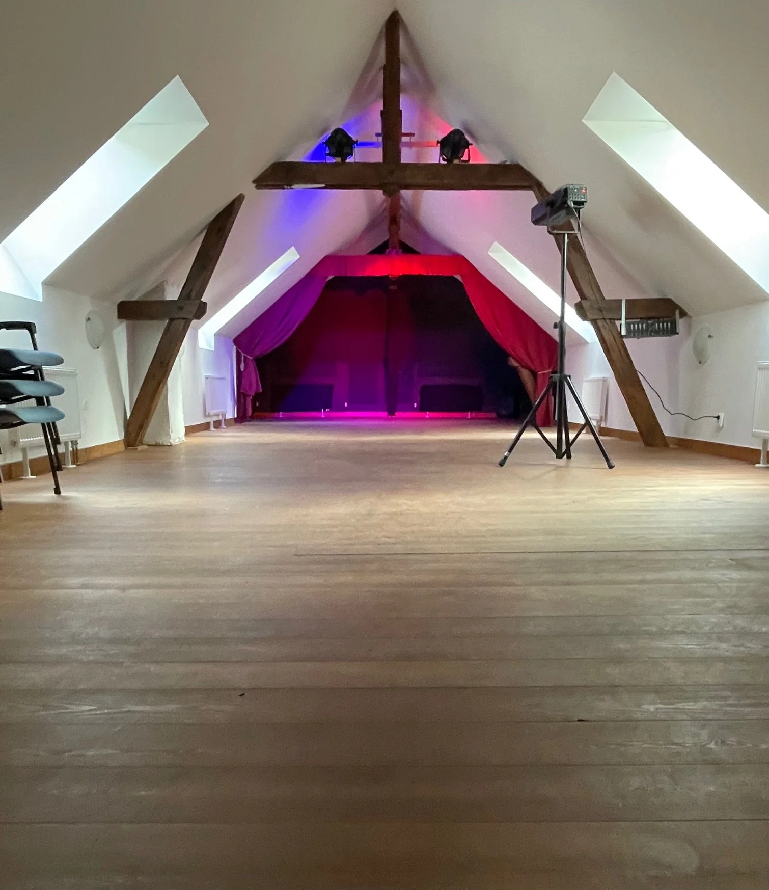 An empty attic room converted into a stage with colorful lighting, a red curtain, wooden beams, and a projector on a stand.