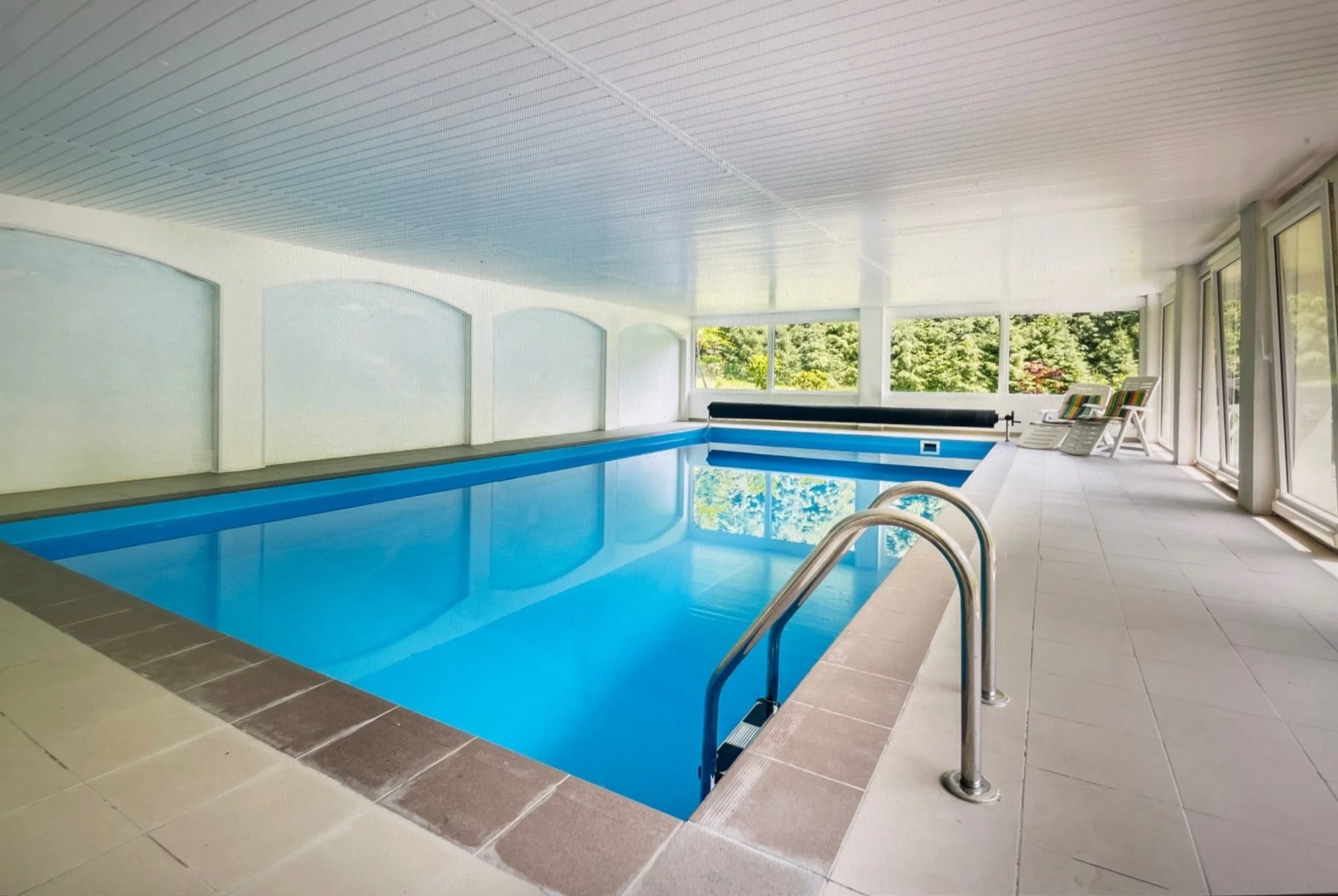 Indoor swimming pool with blue water, white tiled floor, large windows, and outdoor view of green trees, with lounge chairs in the corner.
