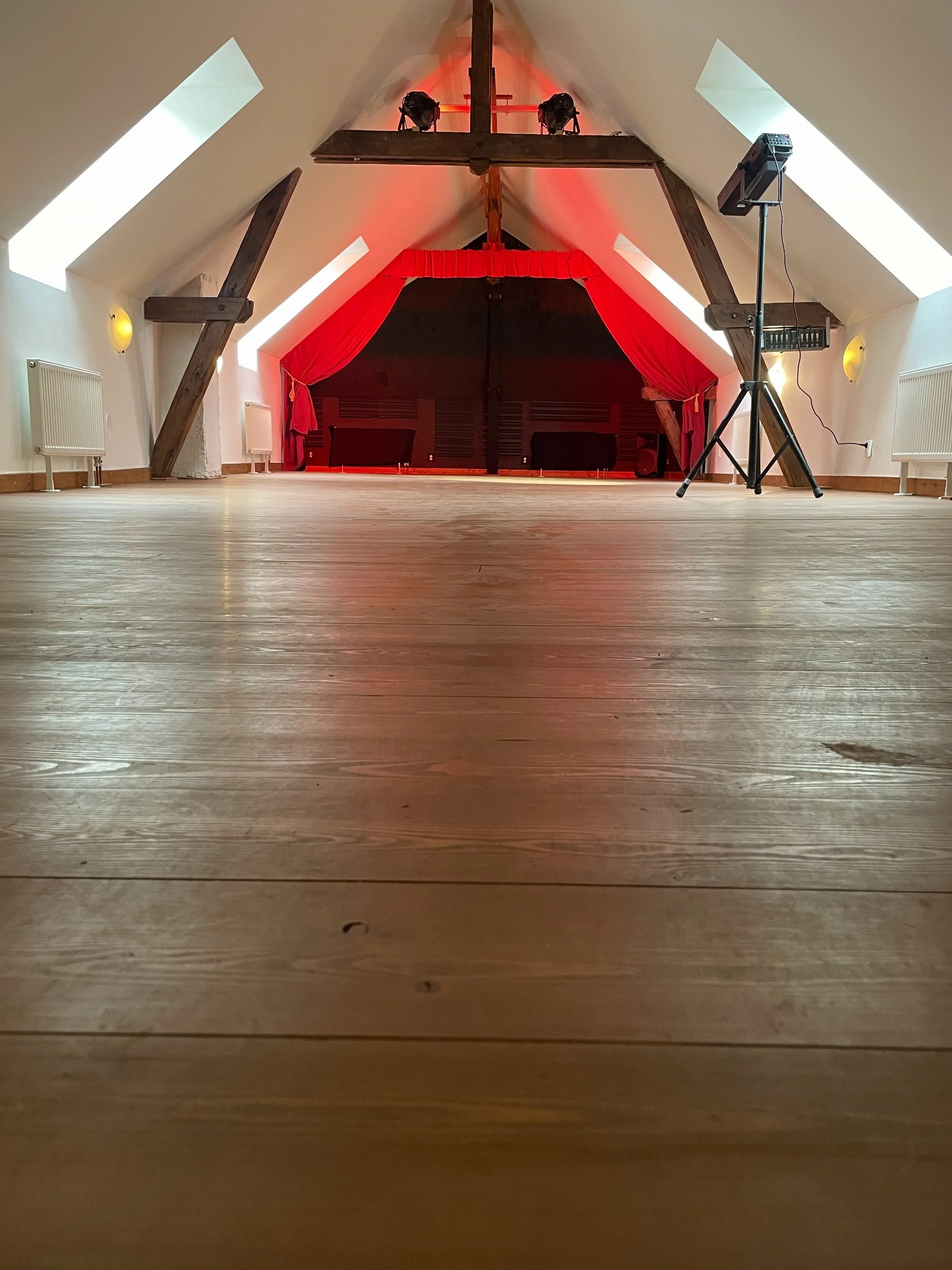 Empty attic room with wooden floor, stage with red curtains, and stage lighting equipment.