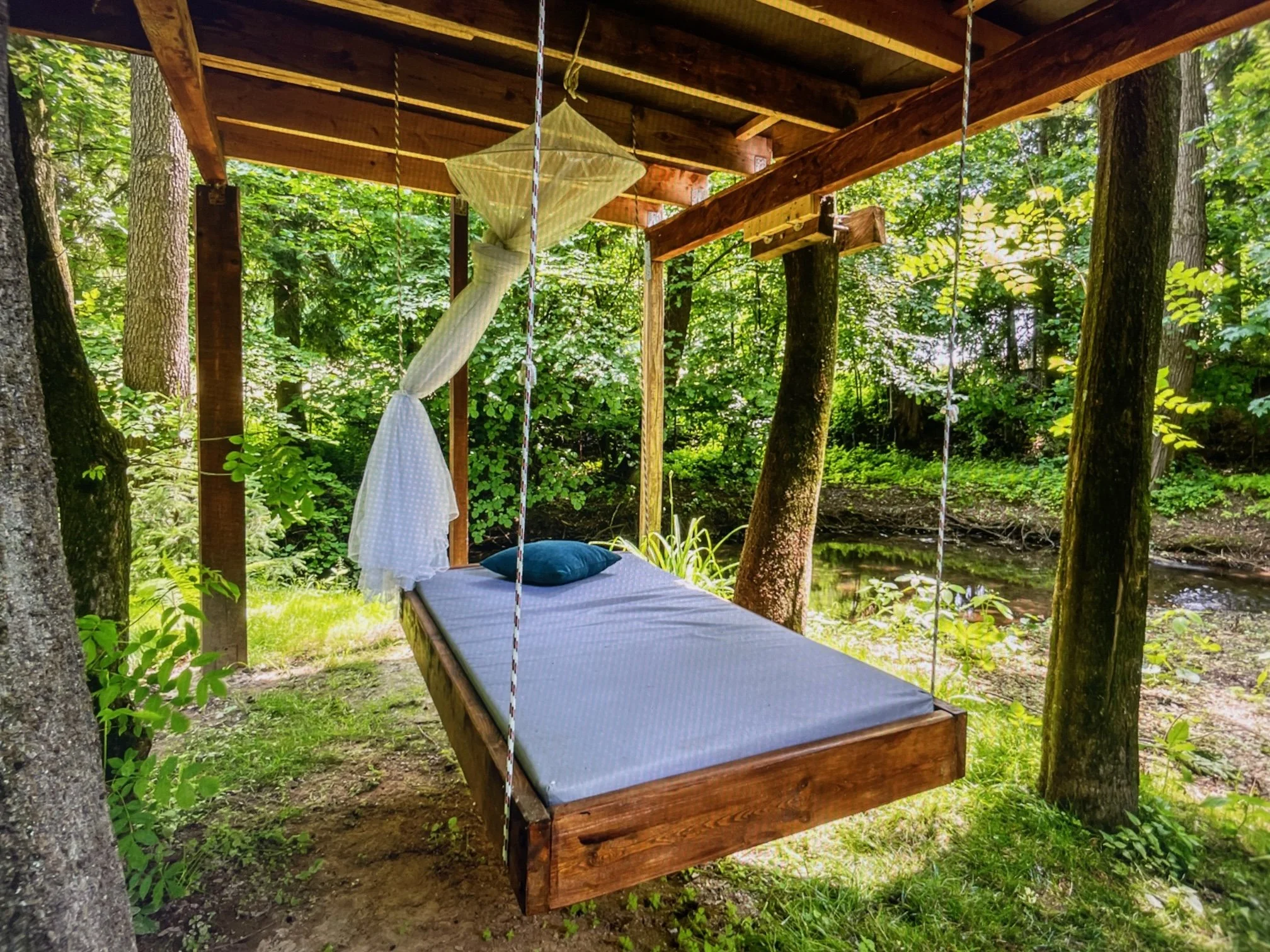 Wooden hammock swing with a mattress, blue pillow, and a white canopy in a forested area near a creek.