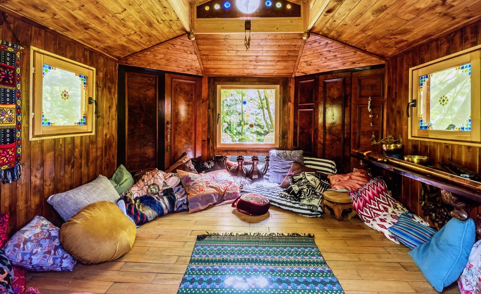 Cozy wooden meditation or yoga room with cushions and pillows, windows with stained glass accents, and a small colorful rug on the wooden floor.