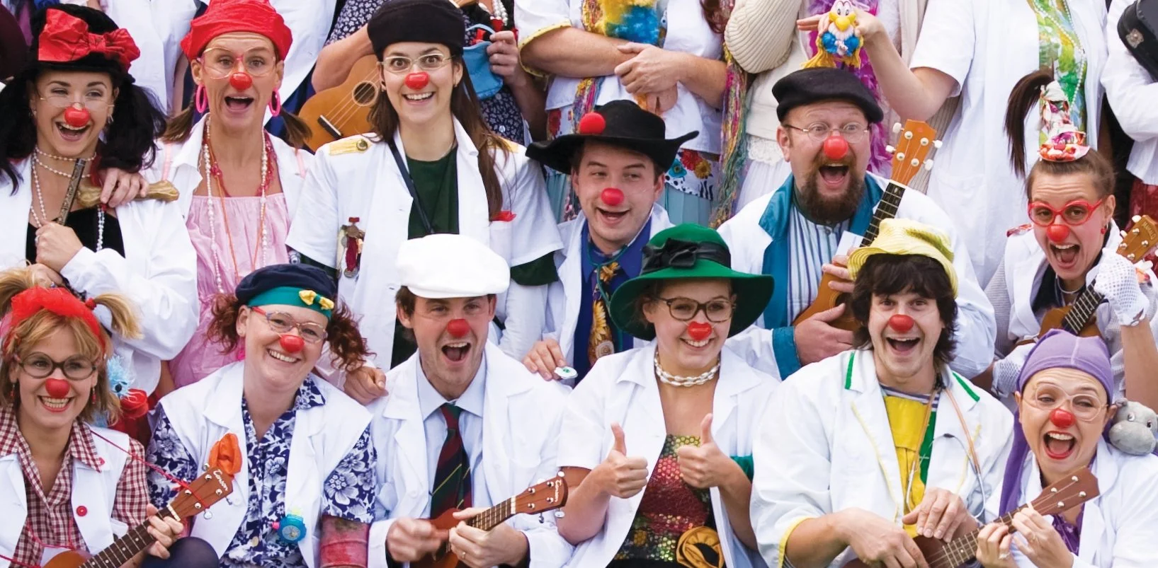 Group of people dressed as clowns with red noses, colorful costumes, and musical instruments, smiling and posing for a photo.