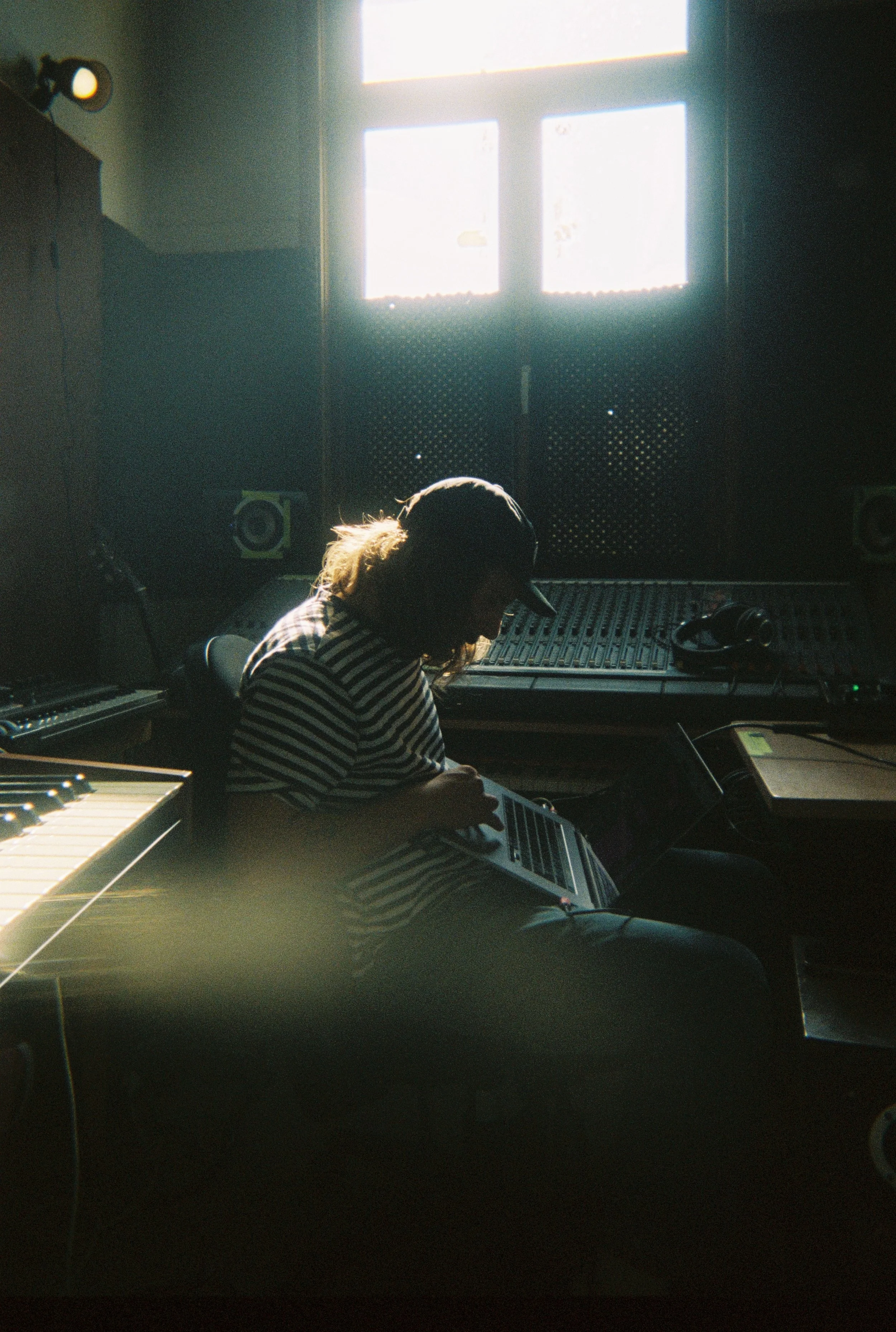 Jelle Rouquart - music producer.  Person with long hair and striped shirt sitting in a dark recording studio, working on music with headphones, illuminated by sunlight through a window.